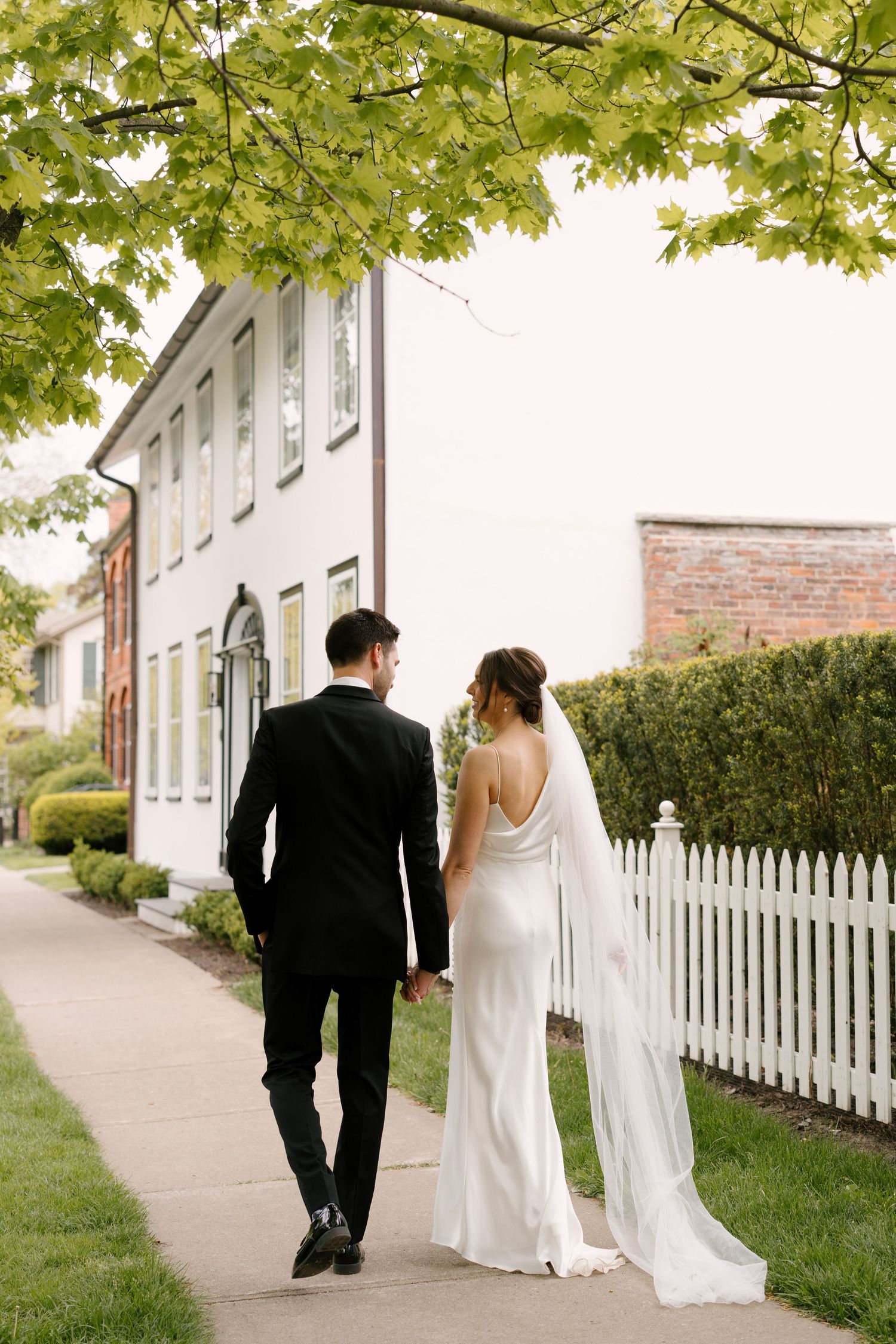 Bride and groom stroll hand in hand past white picket fence and historic building under green trees in Niagara-on-the-lake.