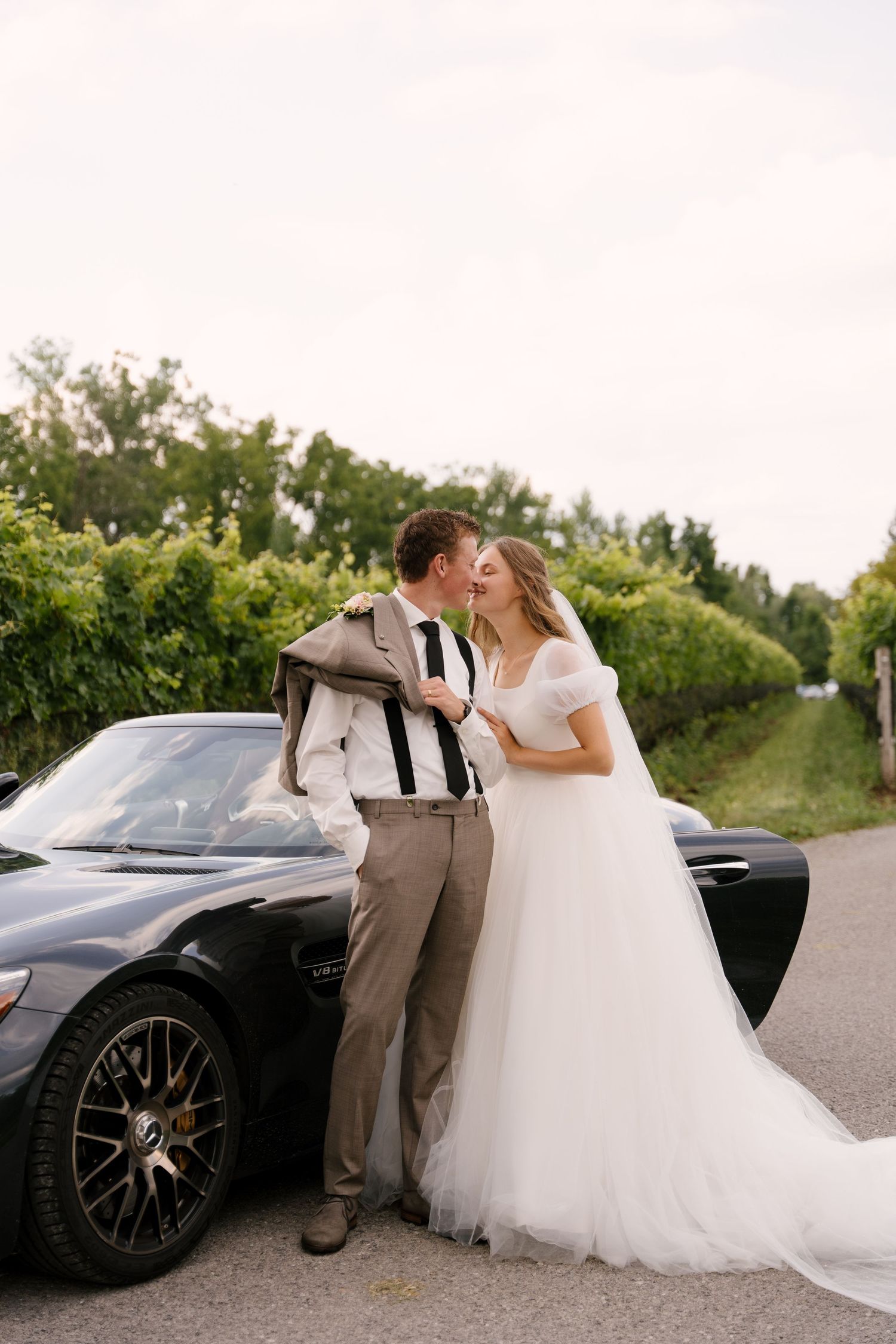 Bride and groom share a kiss leaning against a black sports car beside a Niagara-on-the-lake vineyard road.