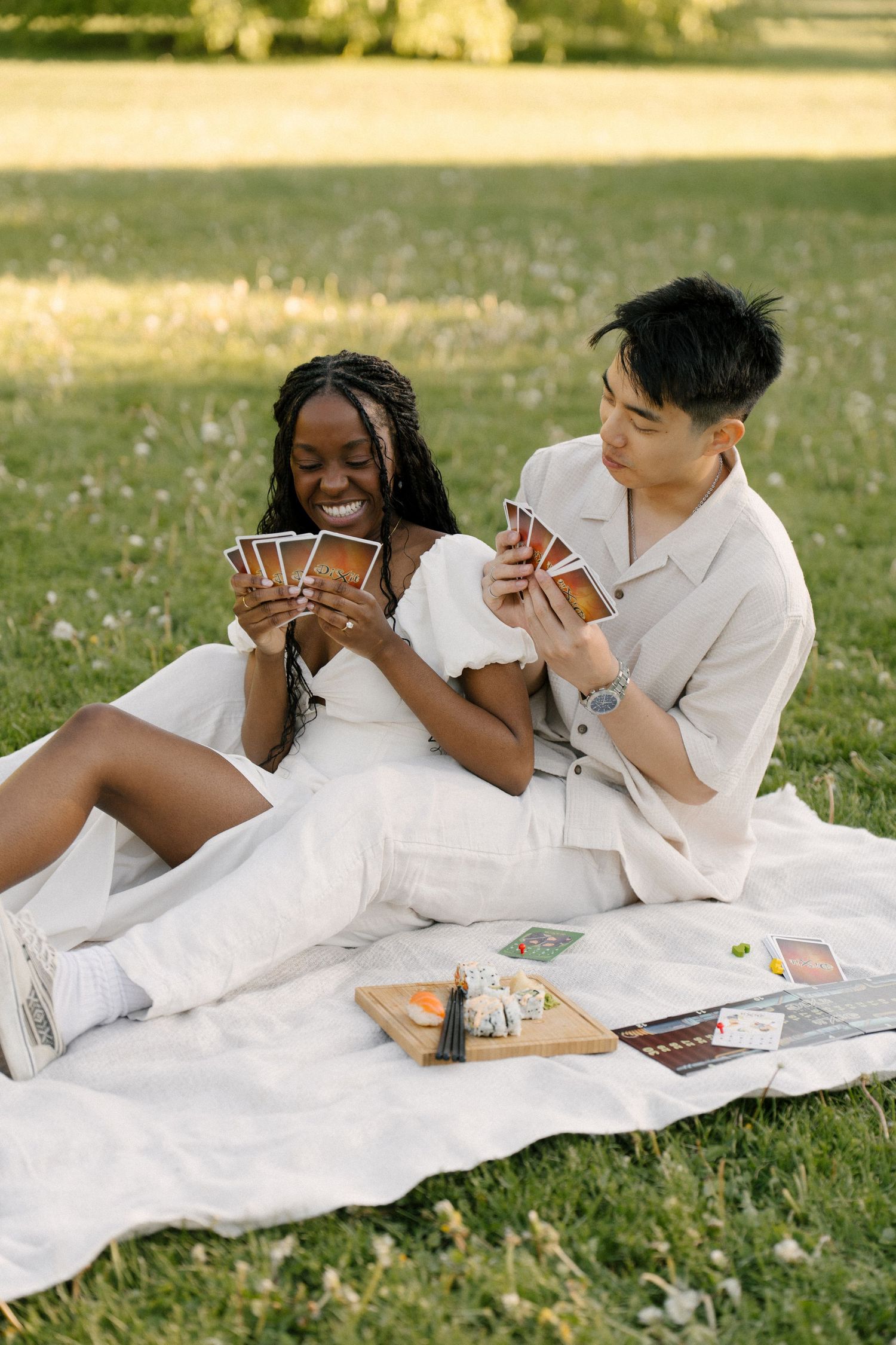 Interracial couple playing cards on a blanket in a sunny park, smiling and enjoying a romantic outdoor picnic.