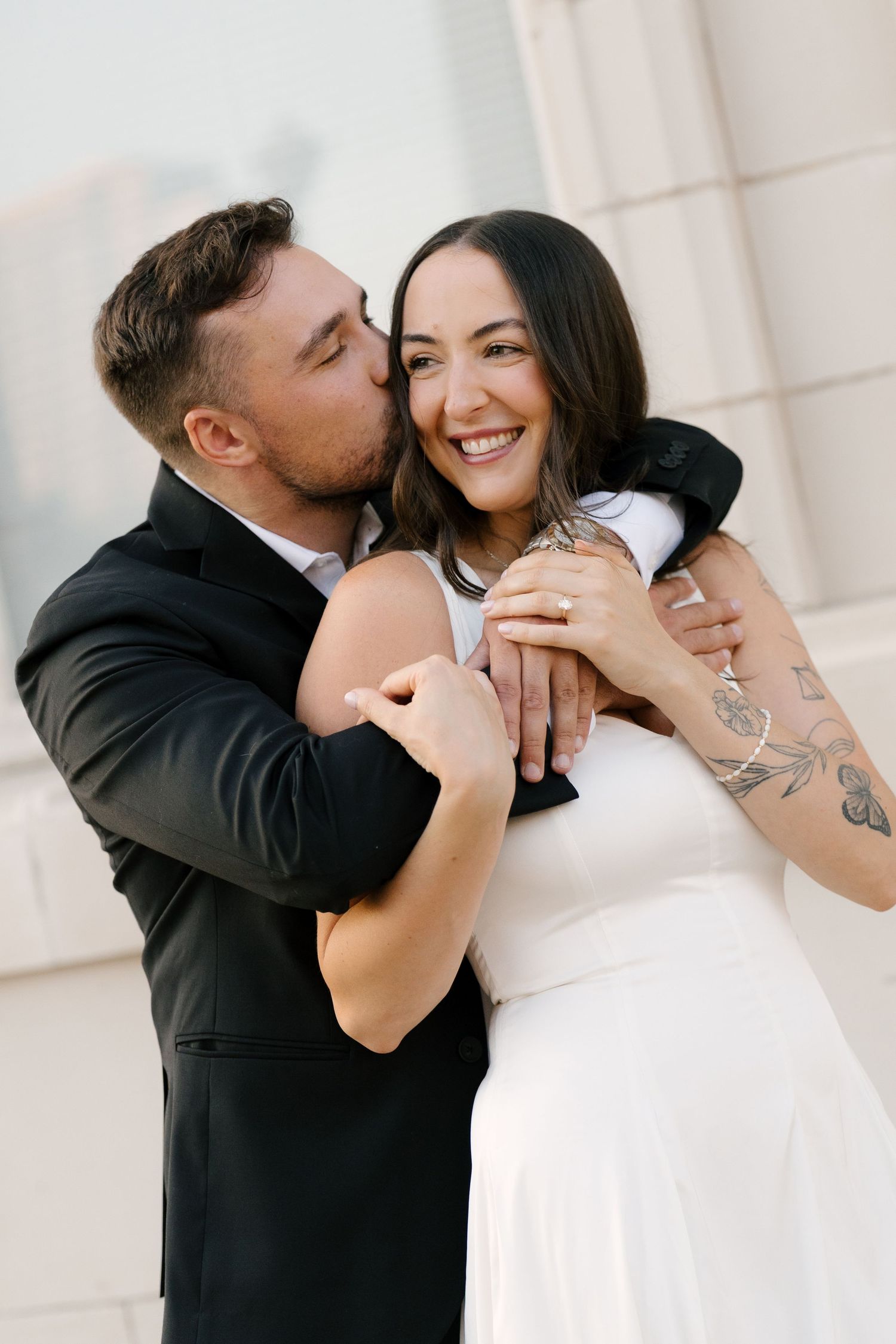 Groom kissing bride on cheek while she smiles, both dressed in wedding attire outdoors.