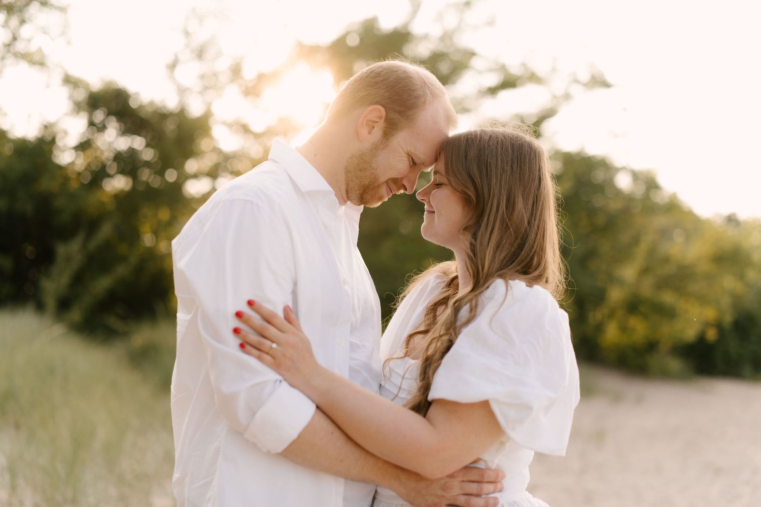 Couple embracing forehead-to-forehead at golden hour on a sandy beach surrounded by green dune grass.