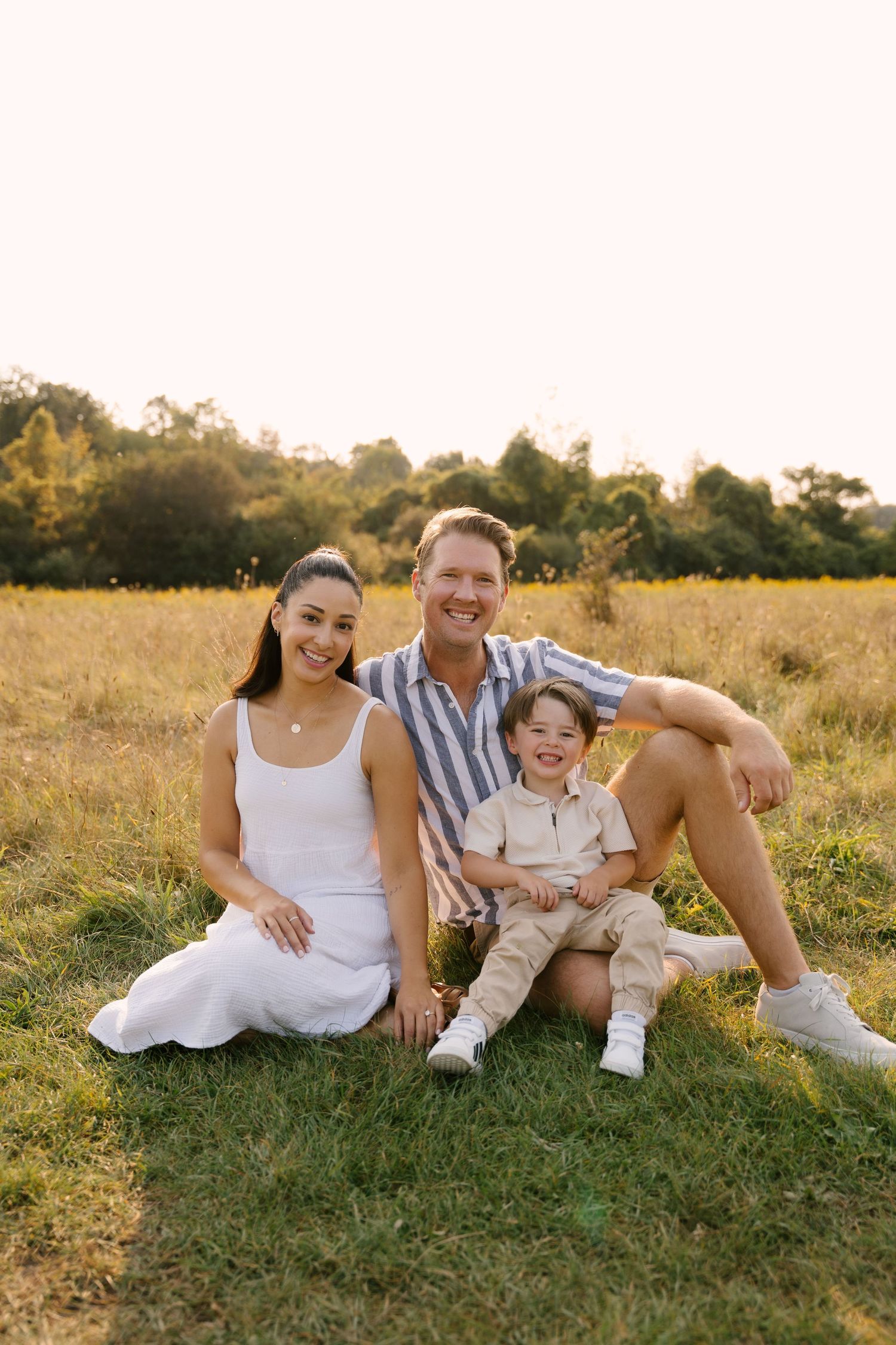 Happy family of three sits together on green grass in a sunny field, smiling at the camera.