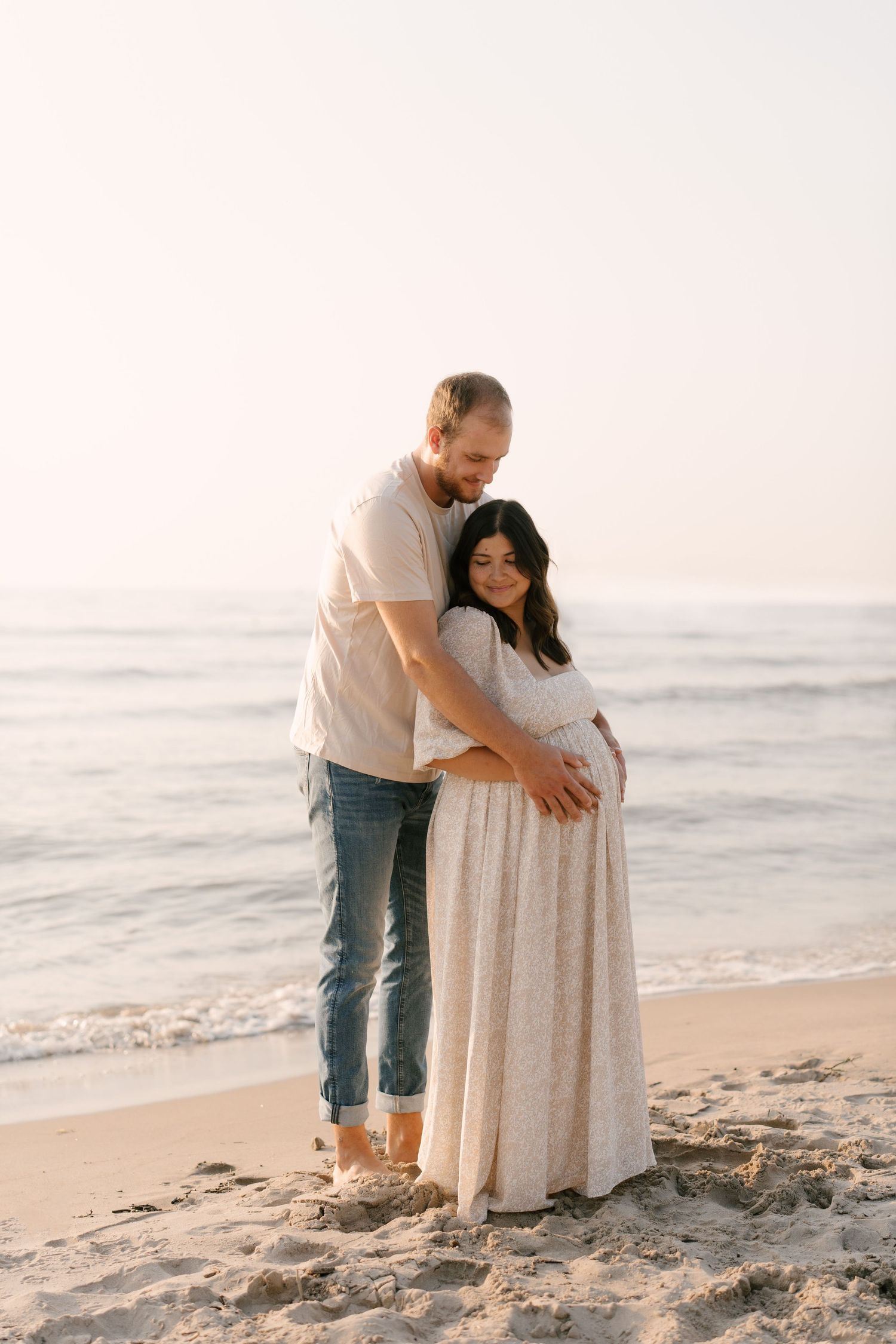 Couple embracing on sandy beach at sunset, pregnant woman in white floral dress held by partner.