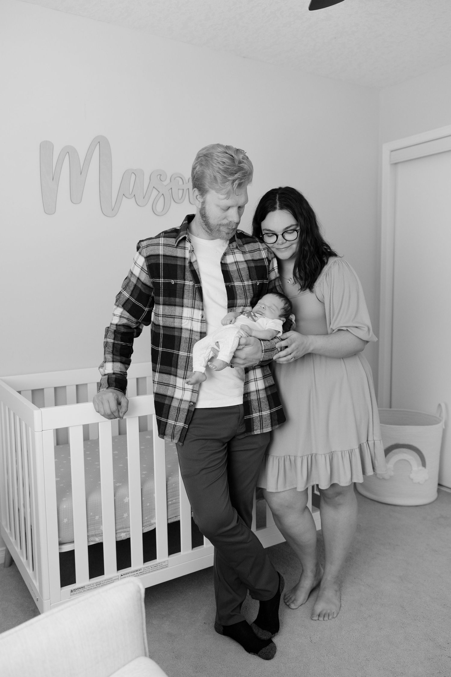 Black and white photo of new parents holding newborn in nursery with Mason name sign.