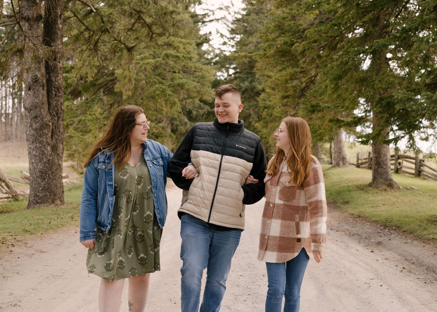 Three young adults laugh and walk together along a tree-lined gravel path during an outdoor photo session.