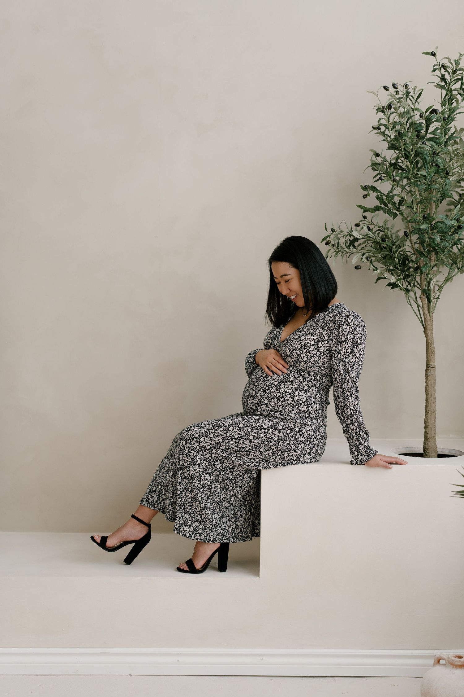Pregnant woman in floral maxi dress sits on white platform, cradling bump, beside an olive tree.