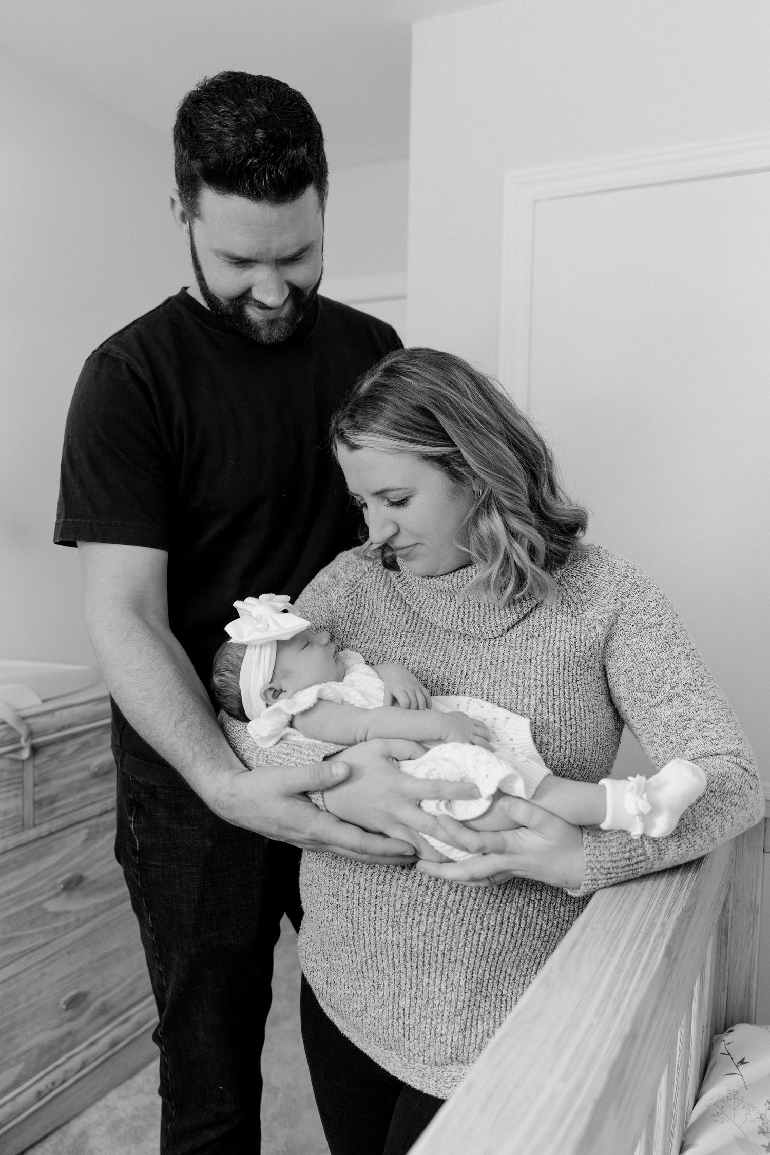 Black and white portrait of new parents lovingly gazing at their newborn baby girl in nursery.