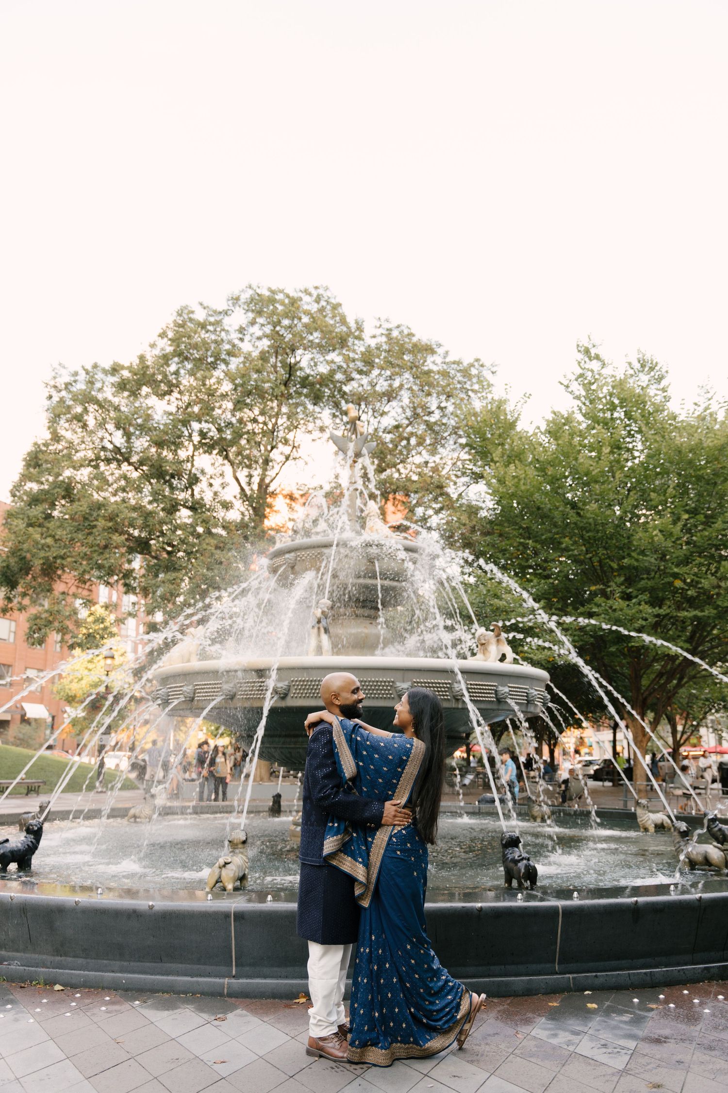 Couple in traditional Indian attire embracing by an ornate fountain surrounded by lush green trees at dusk.