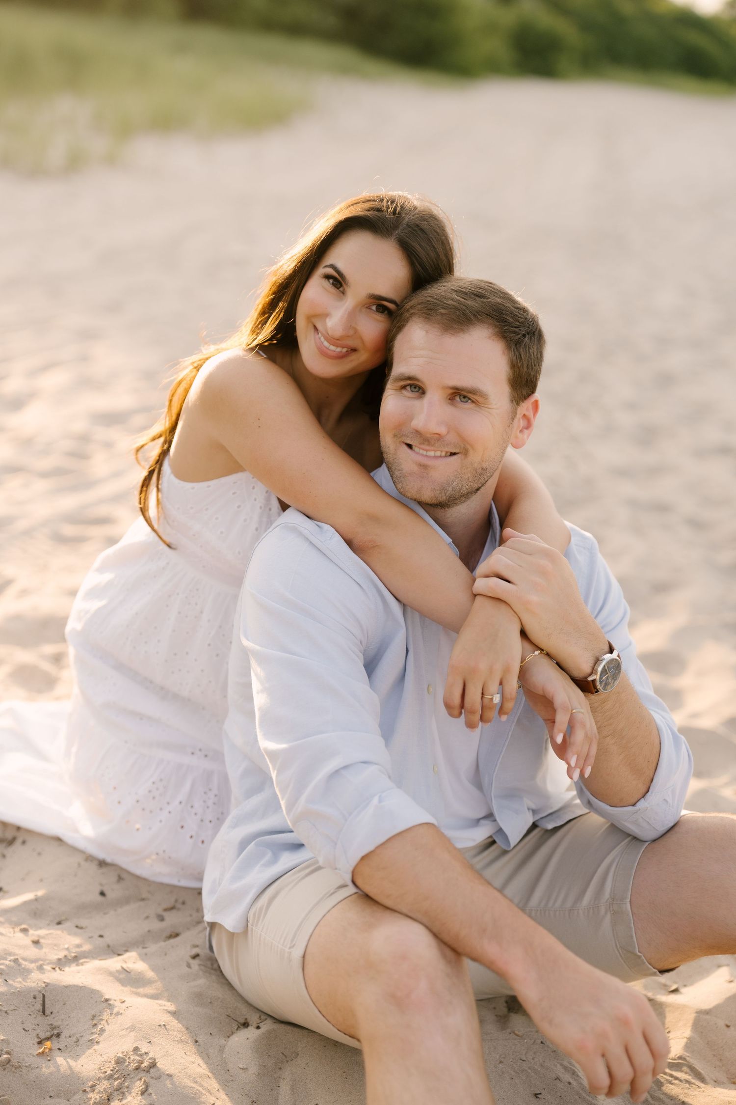 Happy couple sitting on sandy beach at sunset, woman in white dress hugging man from behind smiling.