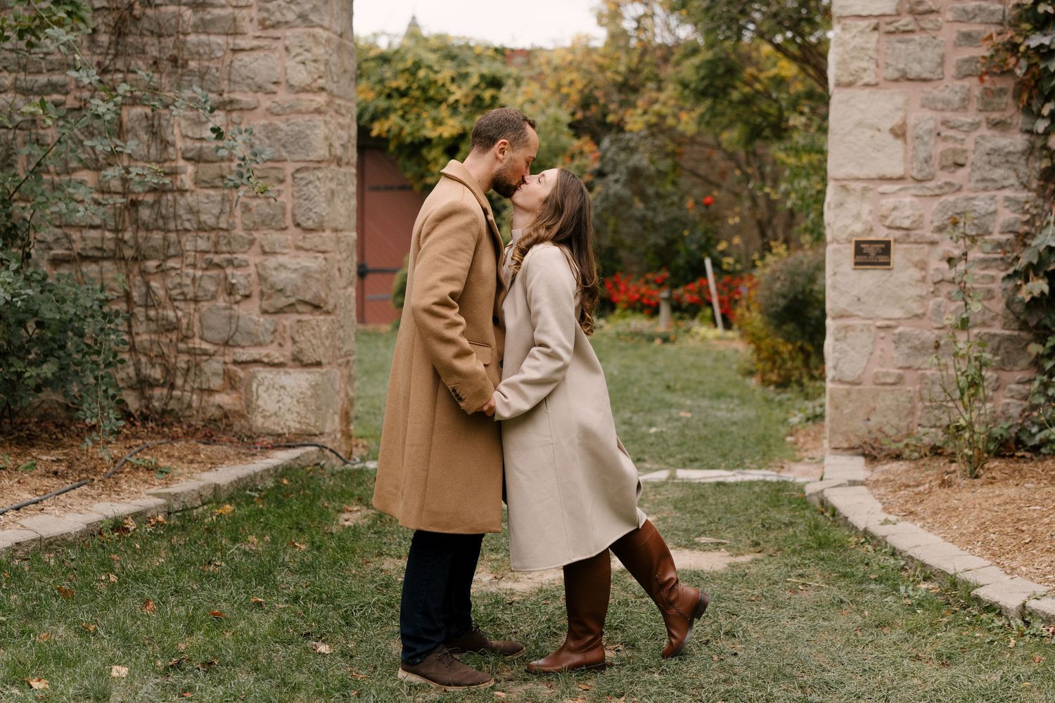 Couple sharing a romantic kiss between stone pillars in an autumn garden, dressed in elegant camel-toned coats.