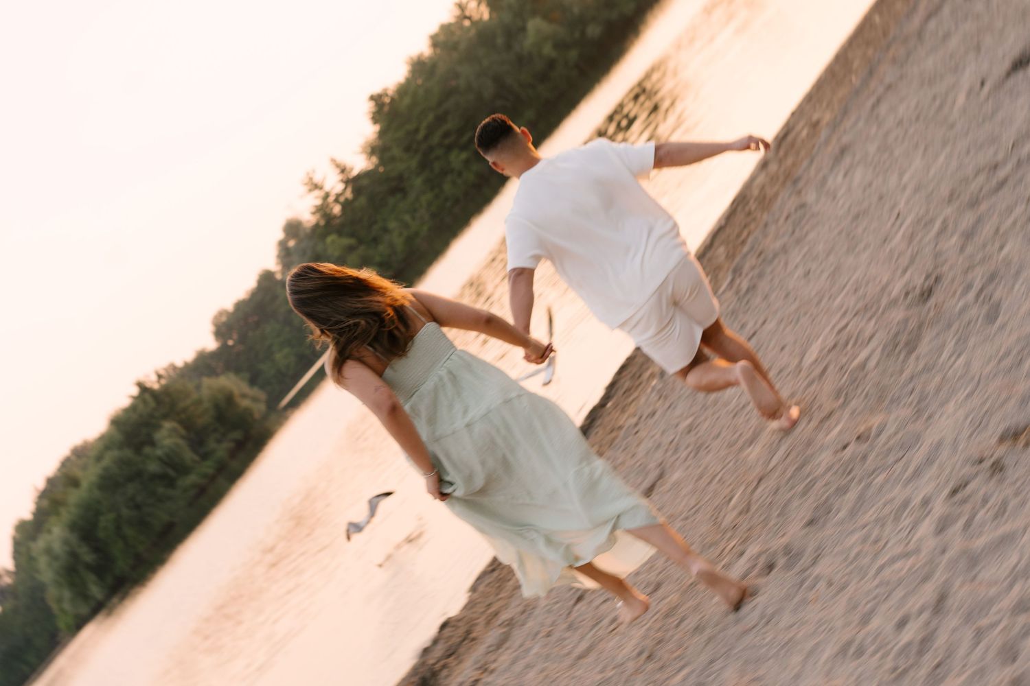 Couple running hand in hand along a sandy beach at sunset with trees in background.
