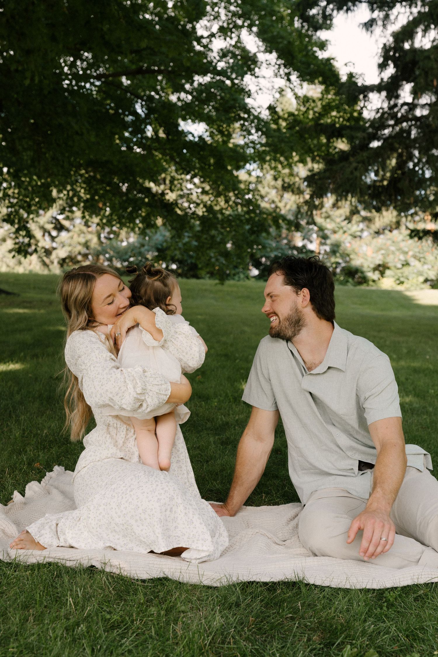 Family sitting on a blanket in a lush green park, mother hugging toddler while father smiles warmly.