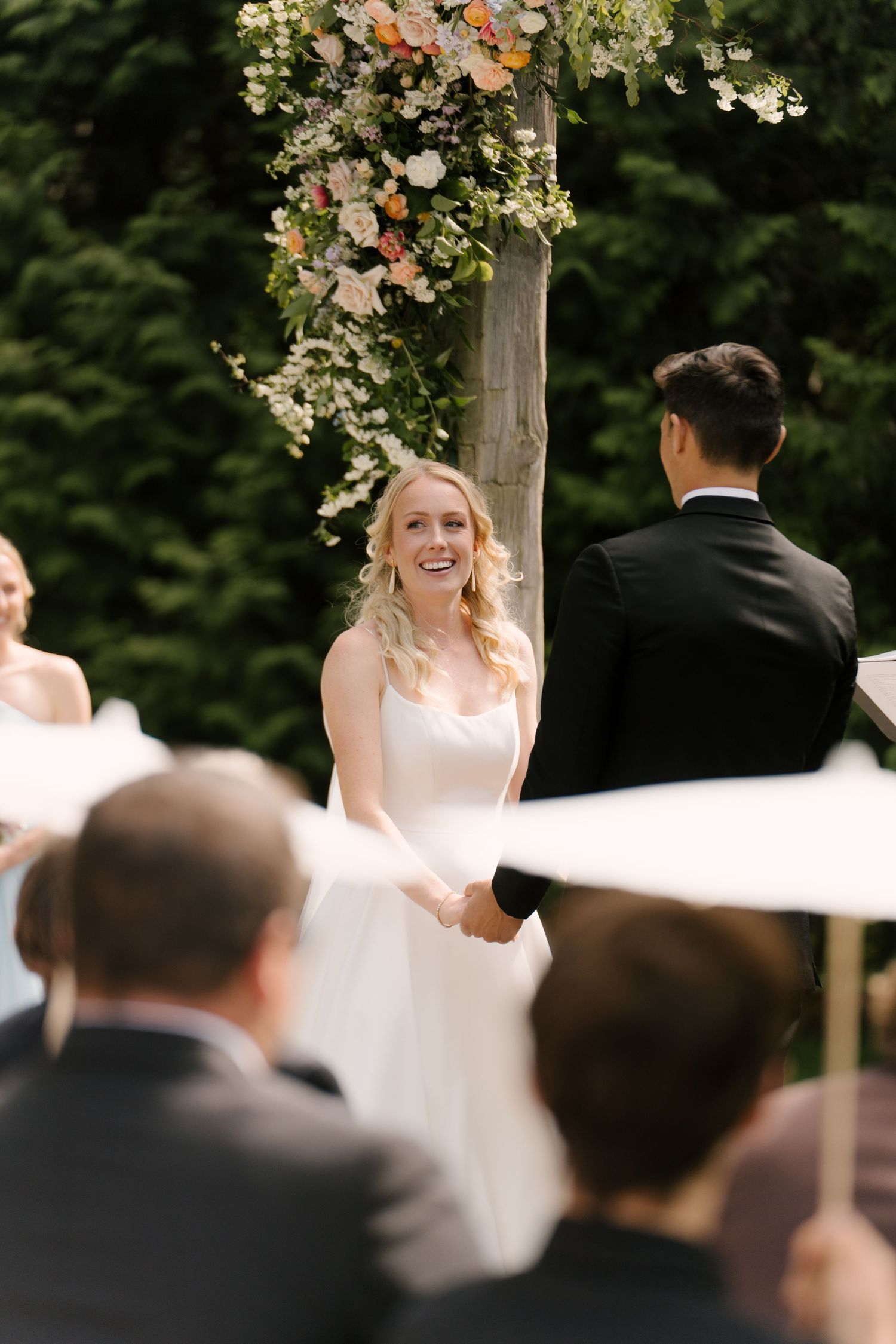 Smiling bride holds grooms hands at outdoor wedding ceremony beneath floral-decorated wooden arch.