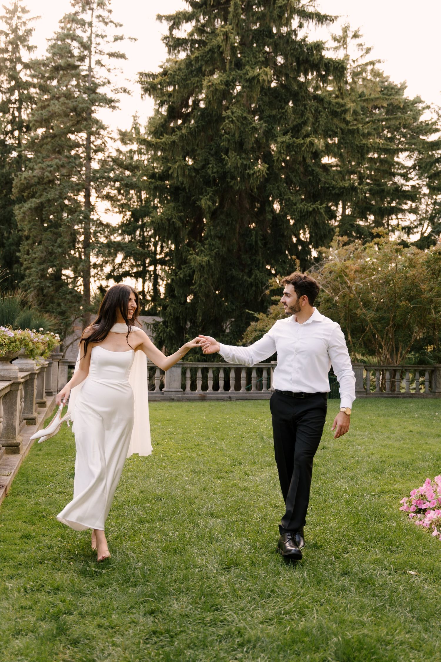 Bride in white gown and groom in white shirt walk hand-in-hand across lush green lawn at estate garden.