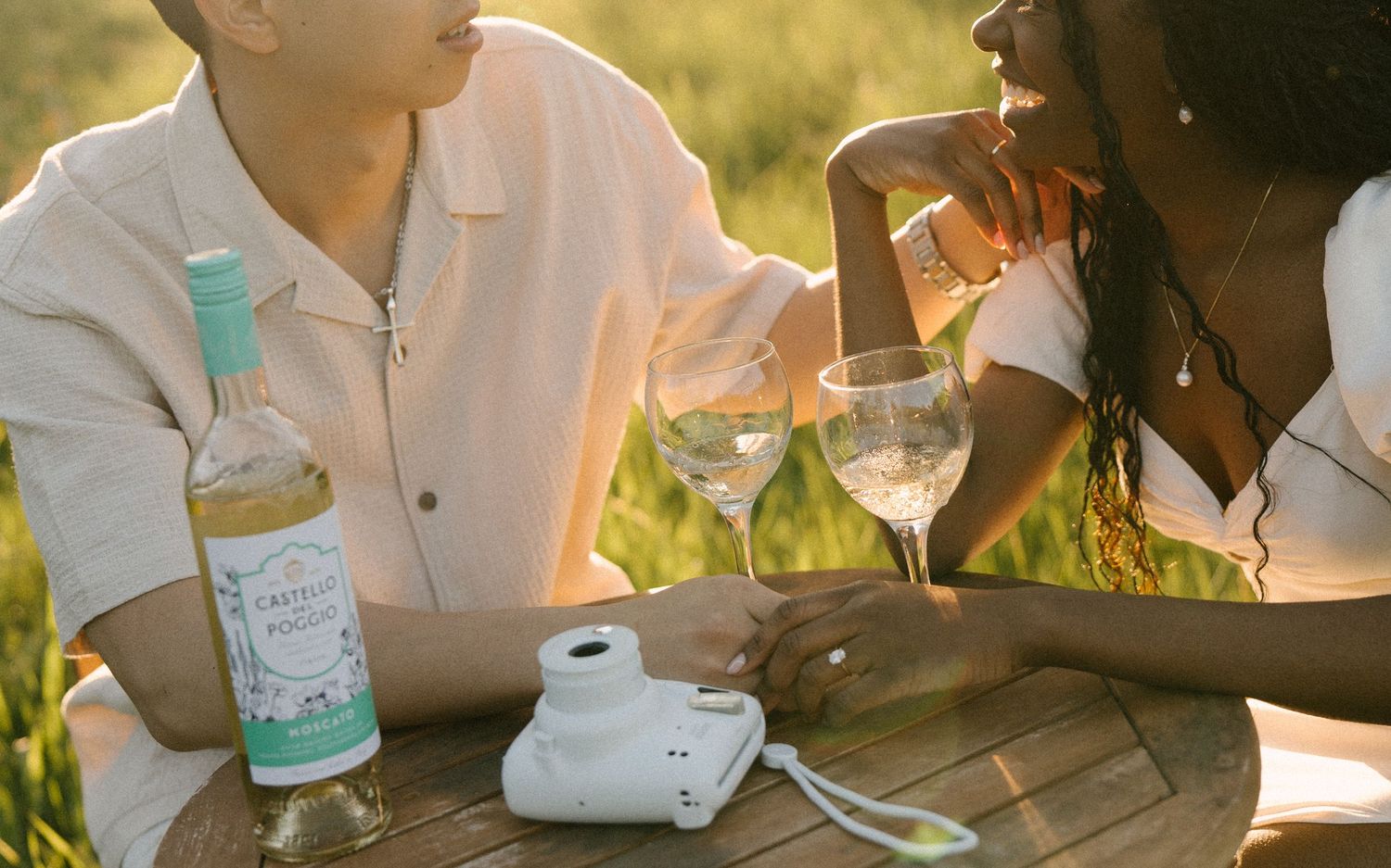 Couple sharing wine with Instax camera on wooden table in sunny meadow.