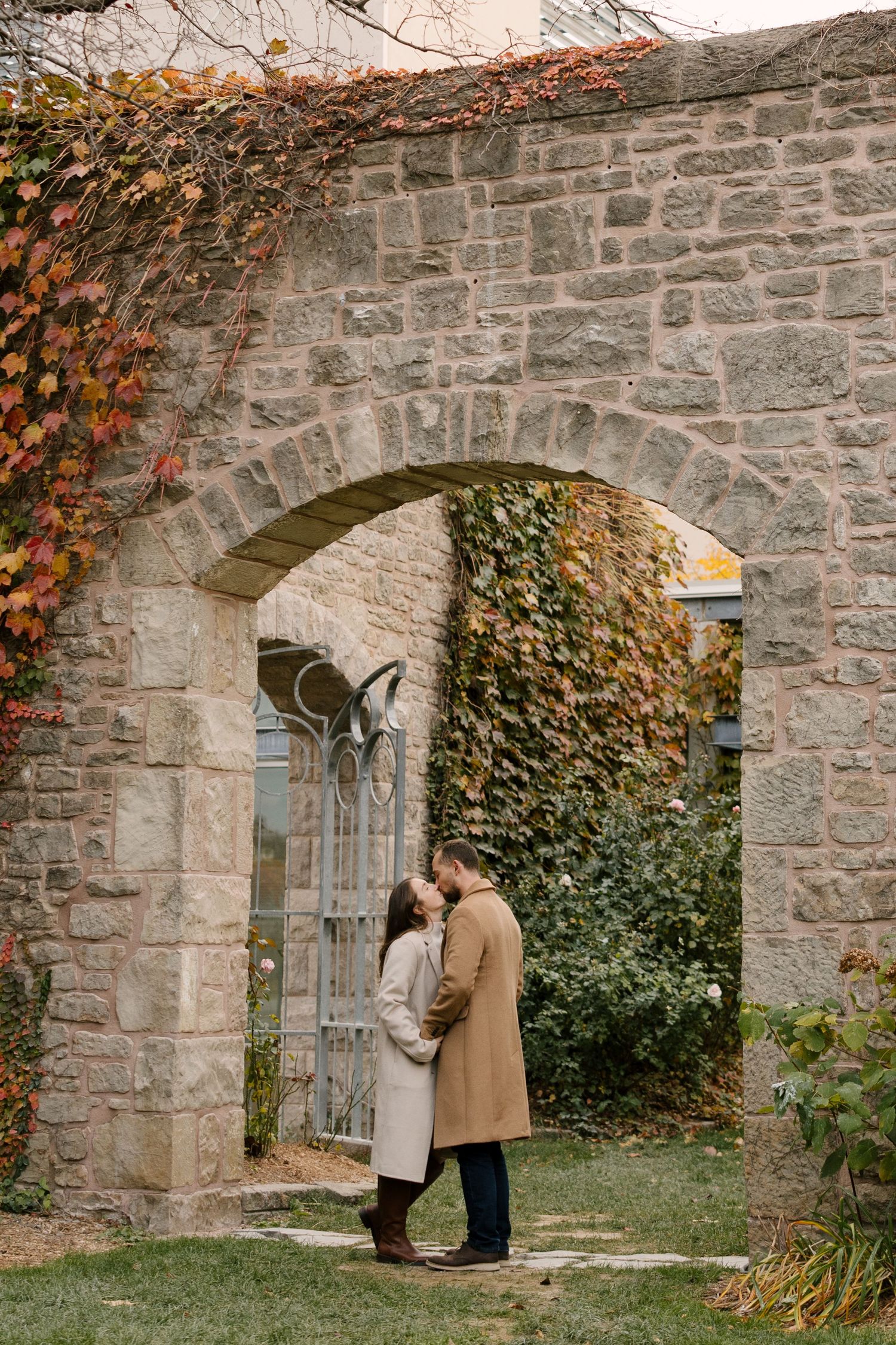Romantic couple sharing a kiss under a vine-covered stone archway in an autumn garden setting.