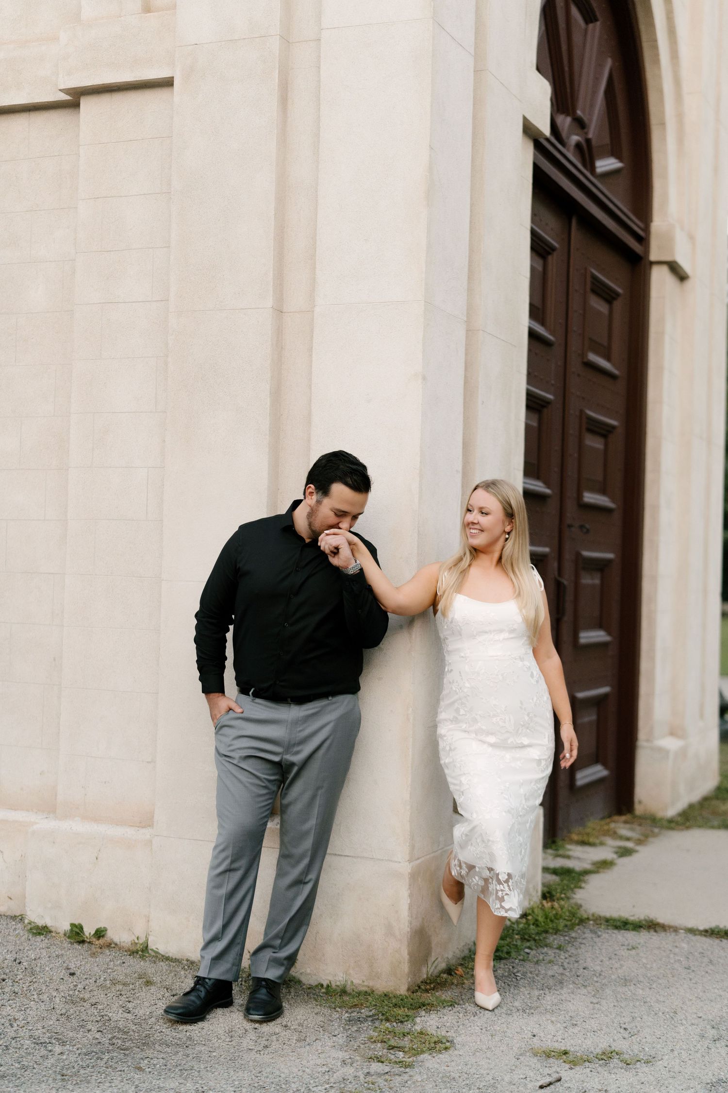 Man in black shirt kissing woman's hand as she smiles against a classic stone building wall.