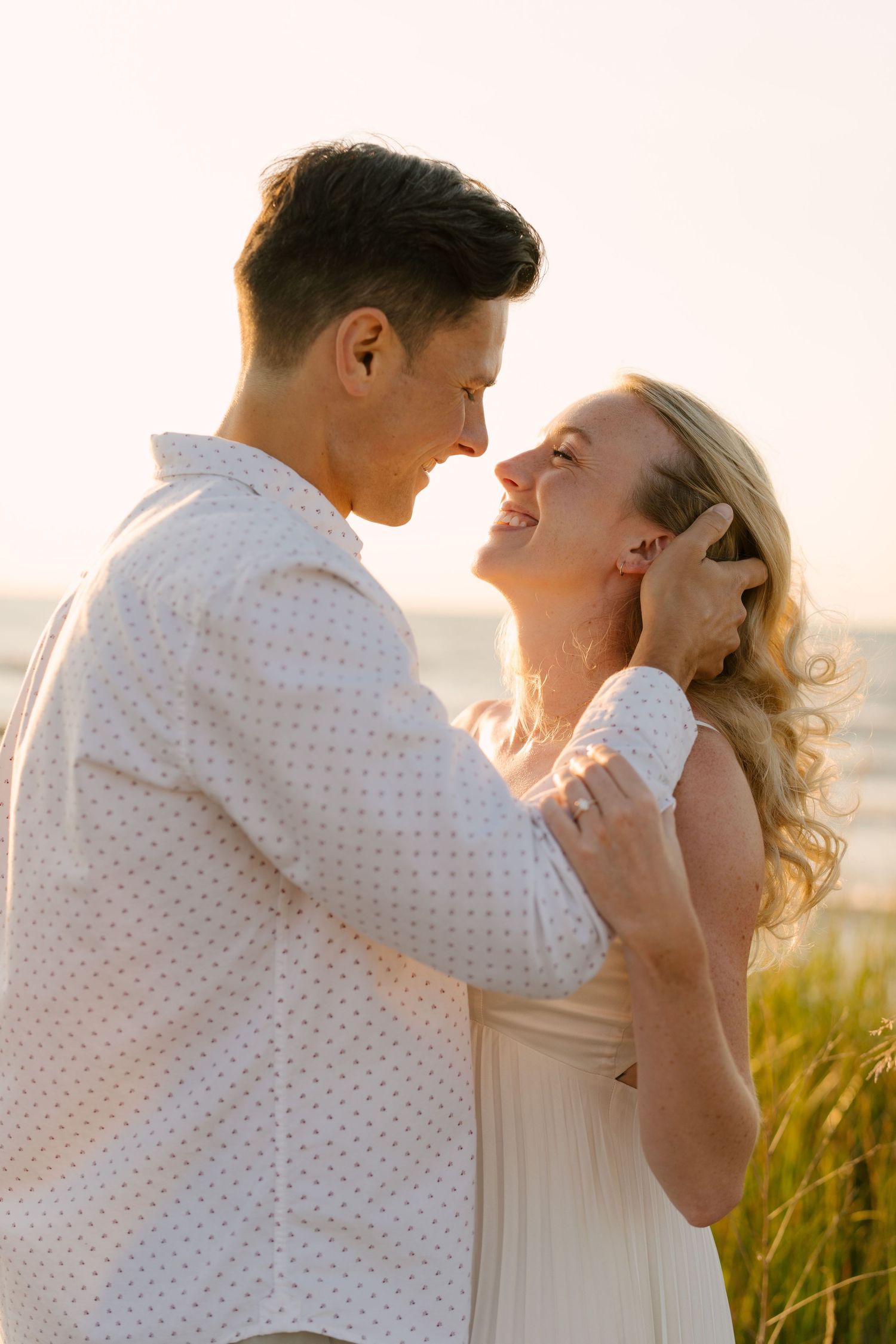 Man tenderly cupping womans face as they lean in close at golden hour near beach grass.