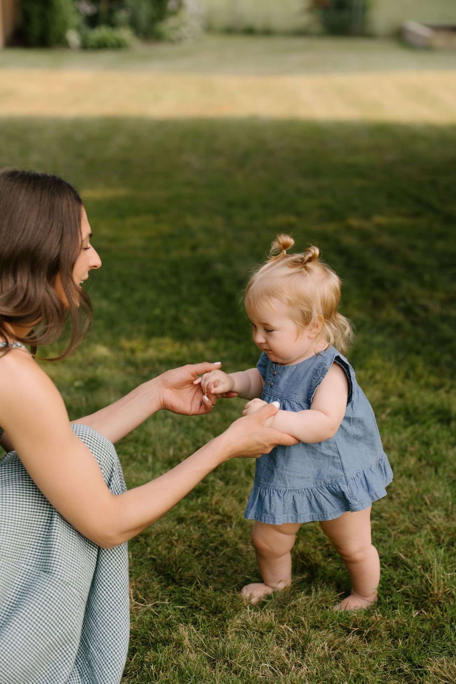 A smiling mother crouches on green grass, holding hands with her curious toddler in a blue dress.
