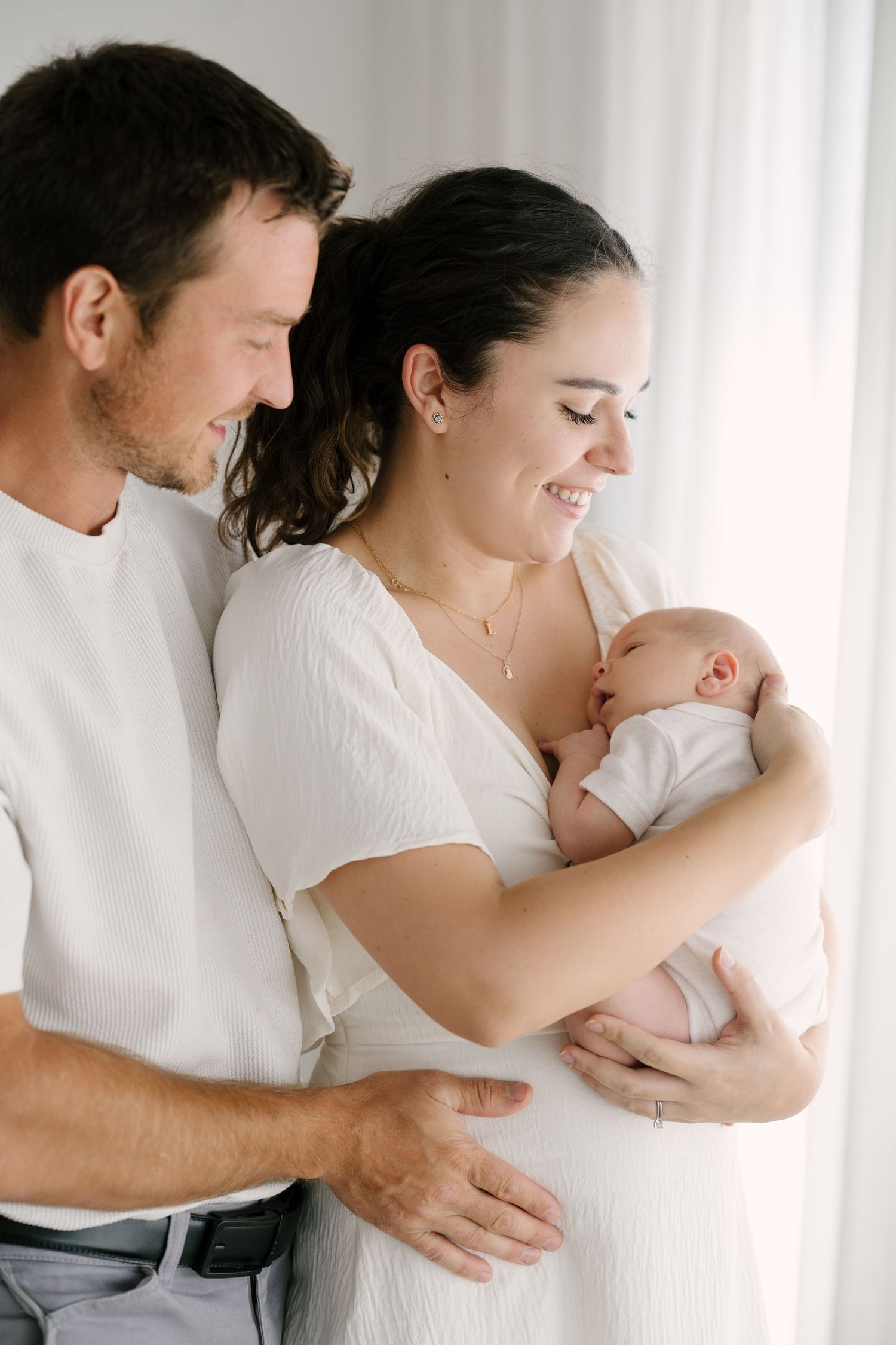 Happy parents in white outfits smiling down at their newborn baby by bright window.
