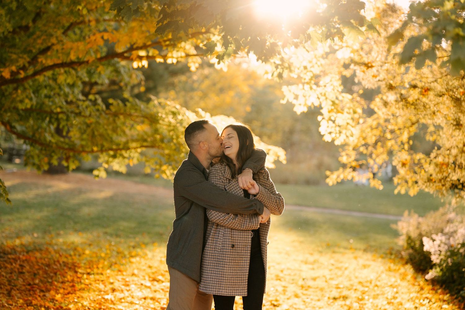 Couple embracing under golden autumn trees in a sunlit park, surrounded by fallen yellow leaves.