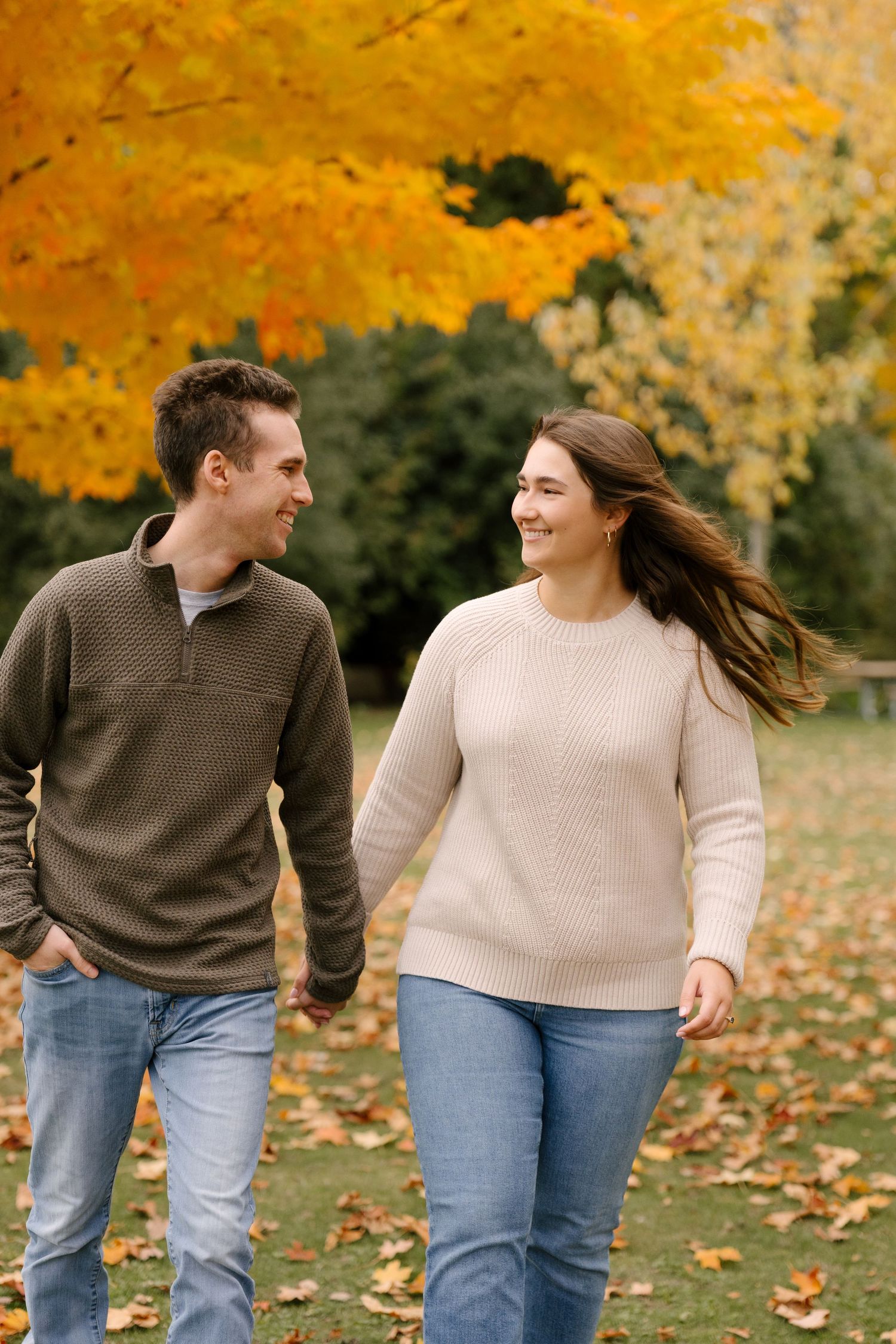 A smiling couple walks hand-in-hand through a park surrounded by vibrant fall foliage.