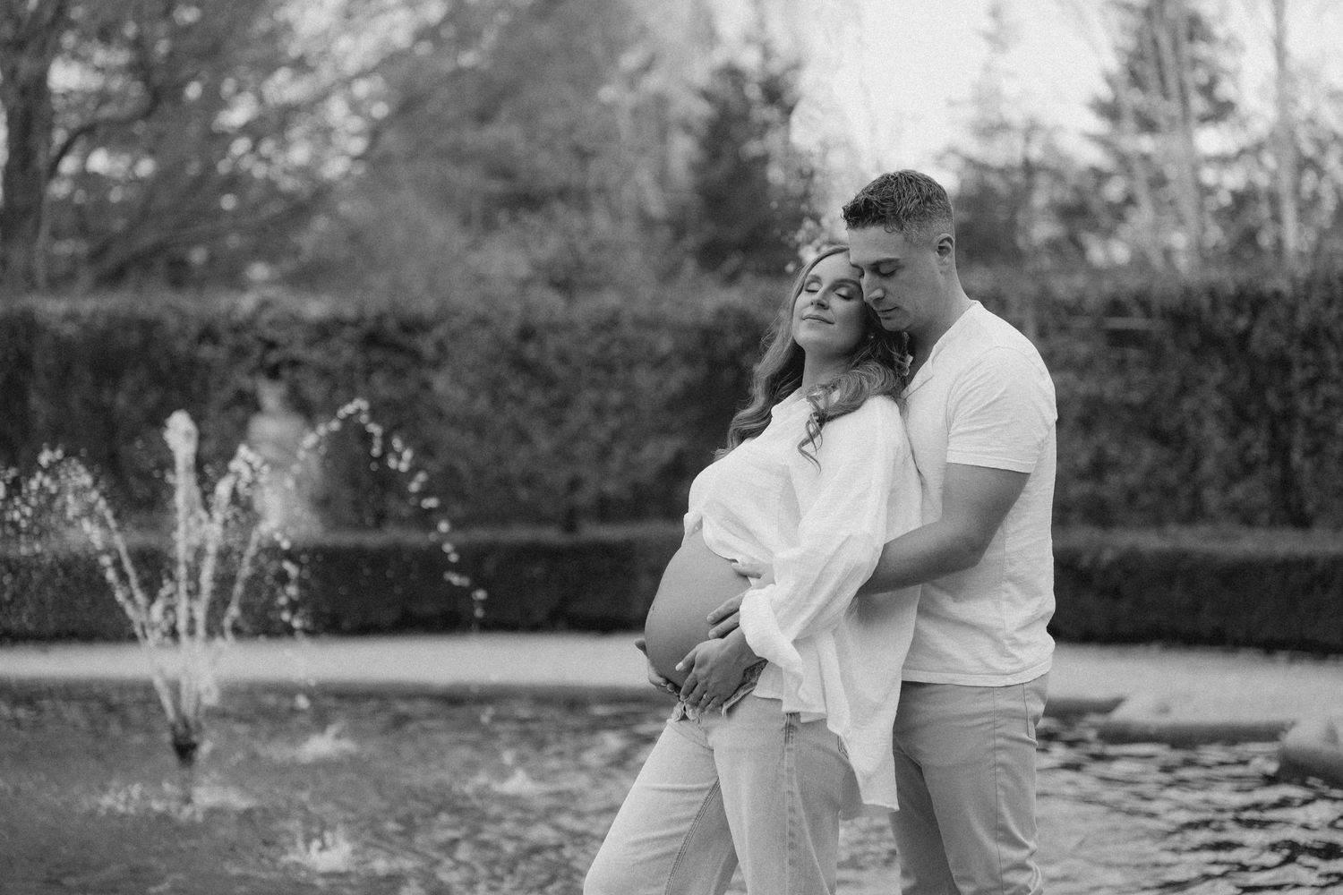 Black and white photo of couple embracing pregnant belly near fountain.