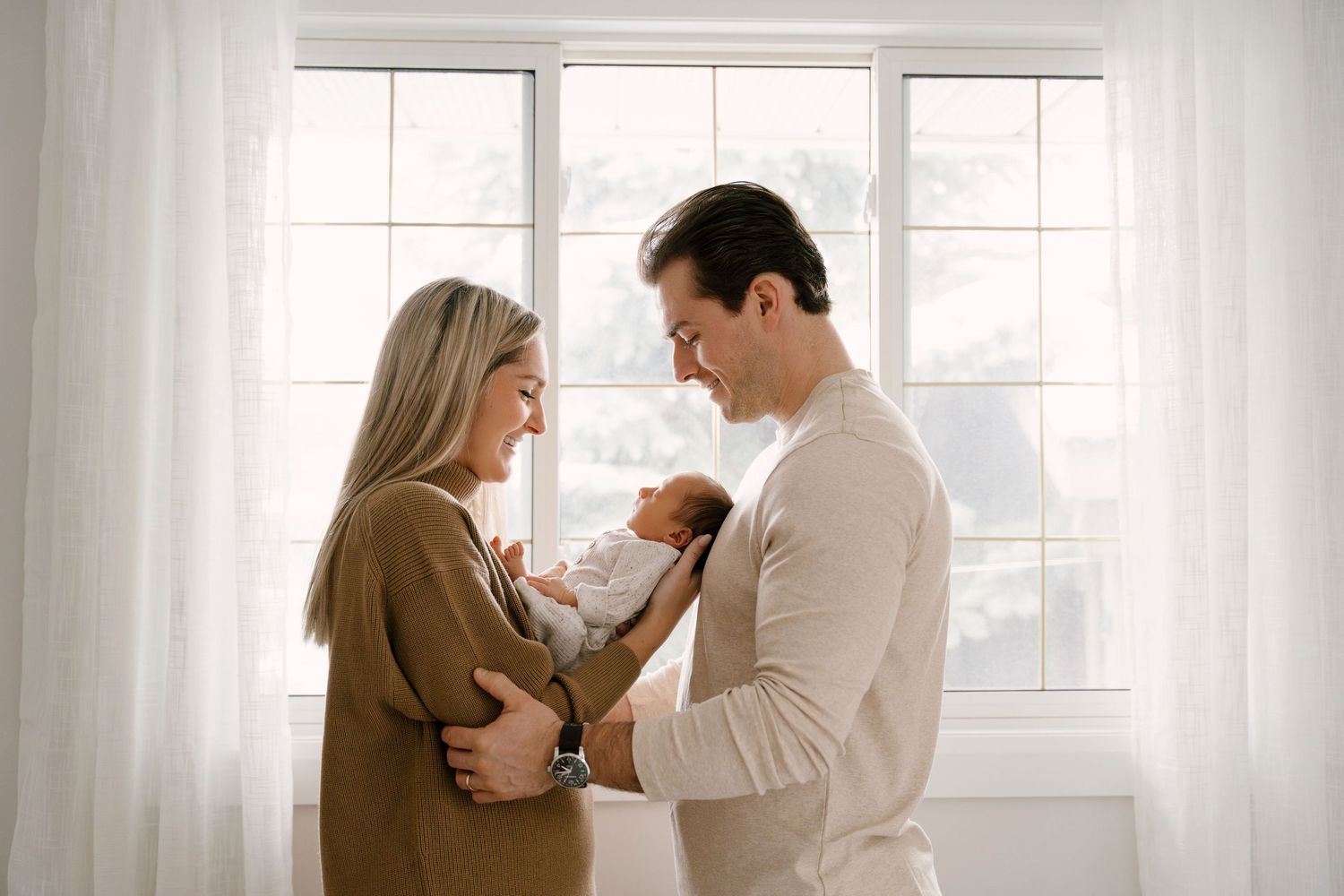 New parents standing by bright window lovingly gazing at their newborn baby wrapped in white.