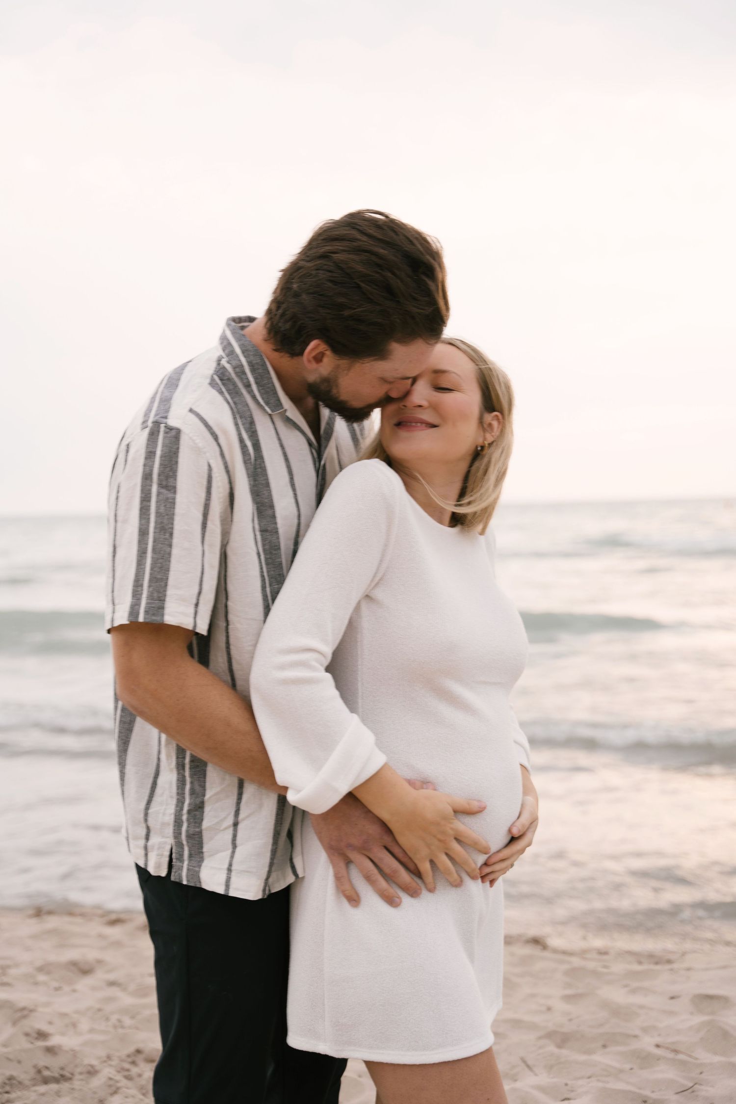Man kissing pregnant partner on cheek while embracing her baby bump on beach.
