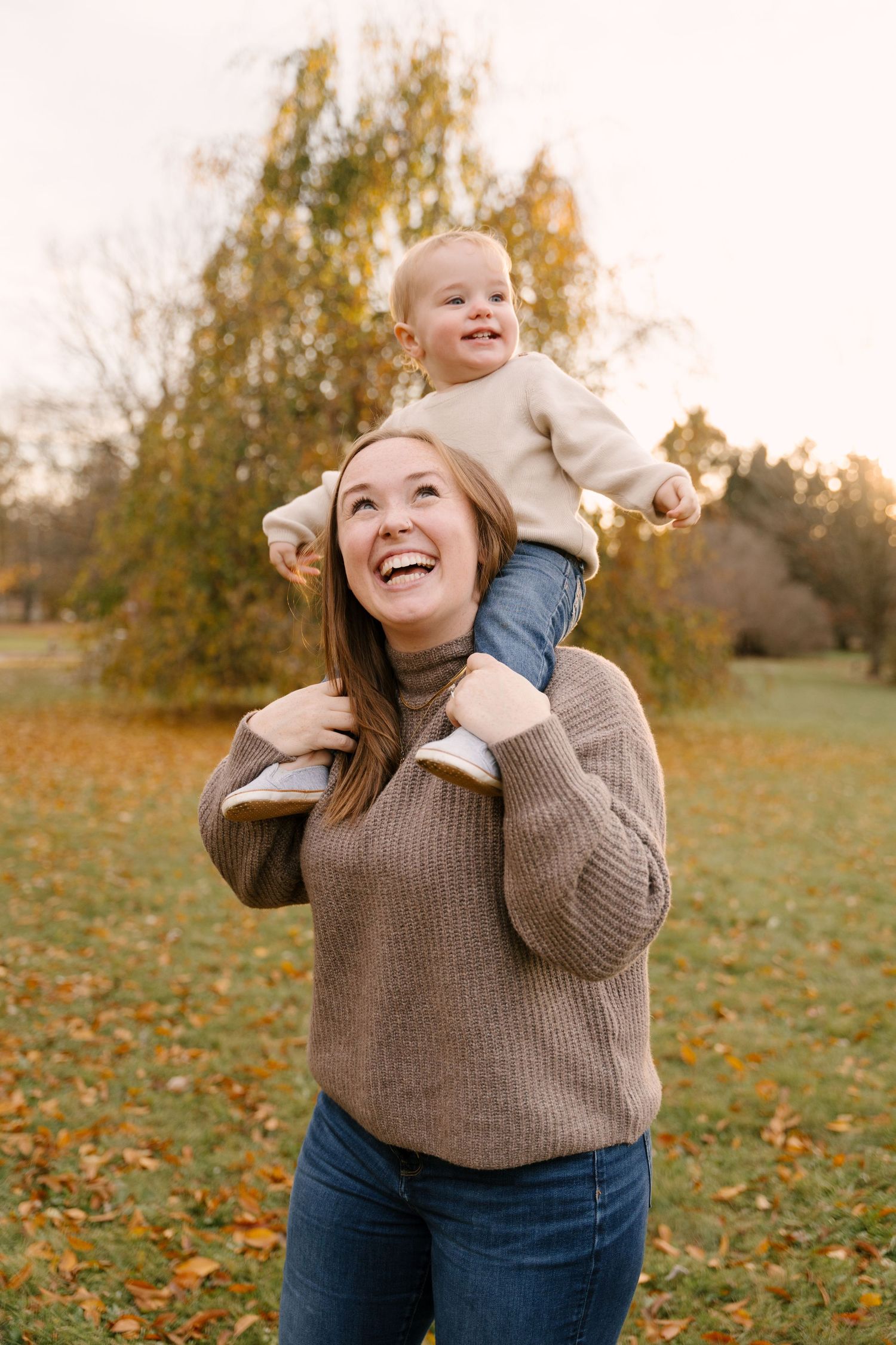 Laughing mother in brown sweater carrying smiling toddler on shoulders in autumn park.