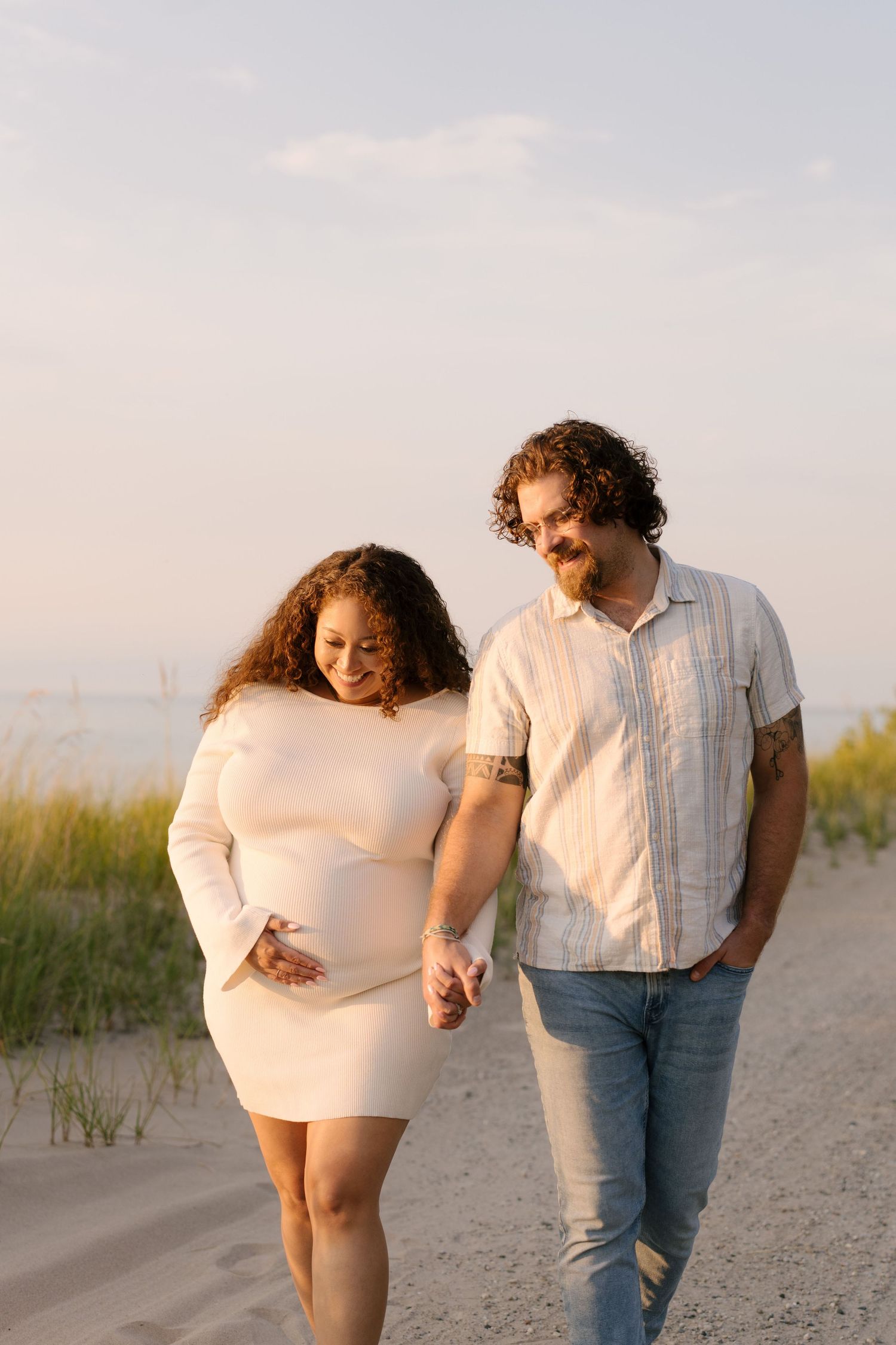 Smiling pregnant couple holding hands and walking along a sandy beach at sunset.