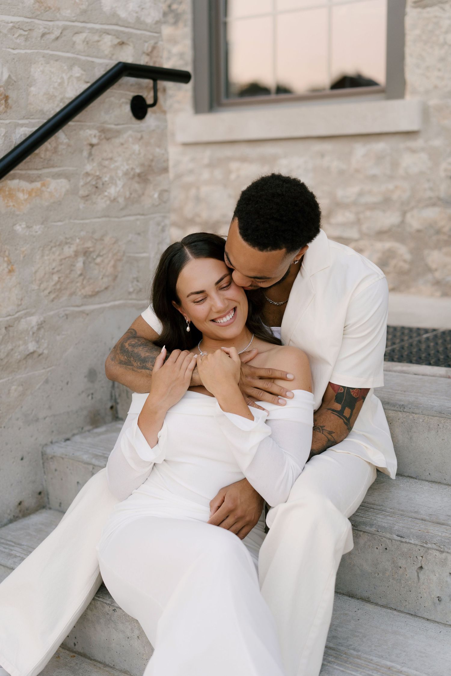 Happy couple in white sit on stone steps, man kisses woman's cheek as she laughs joyfully.