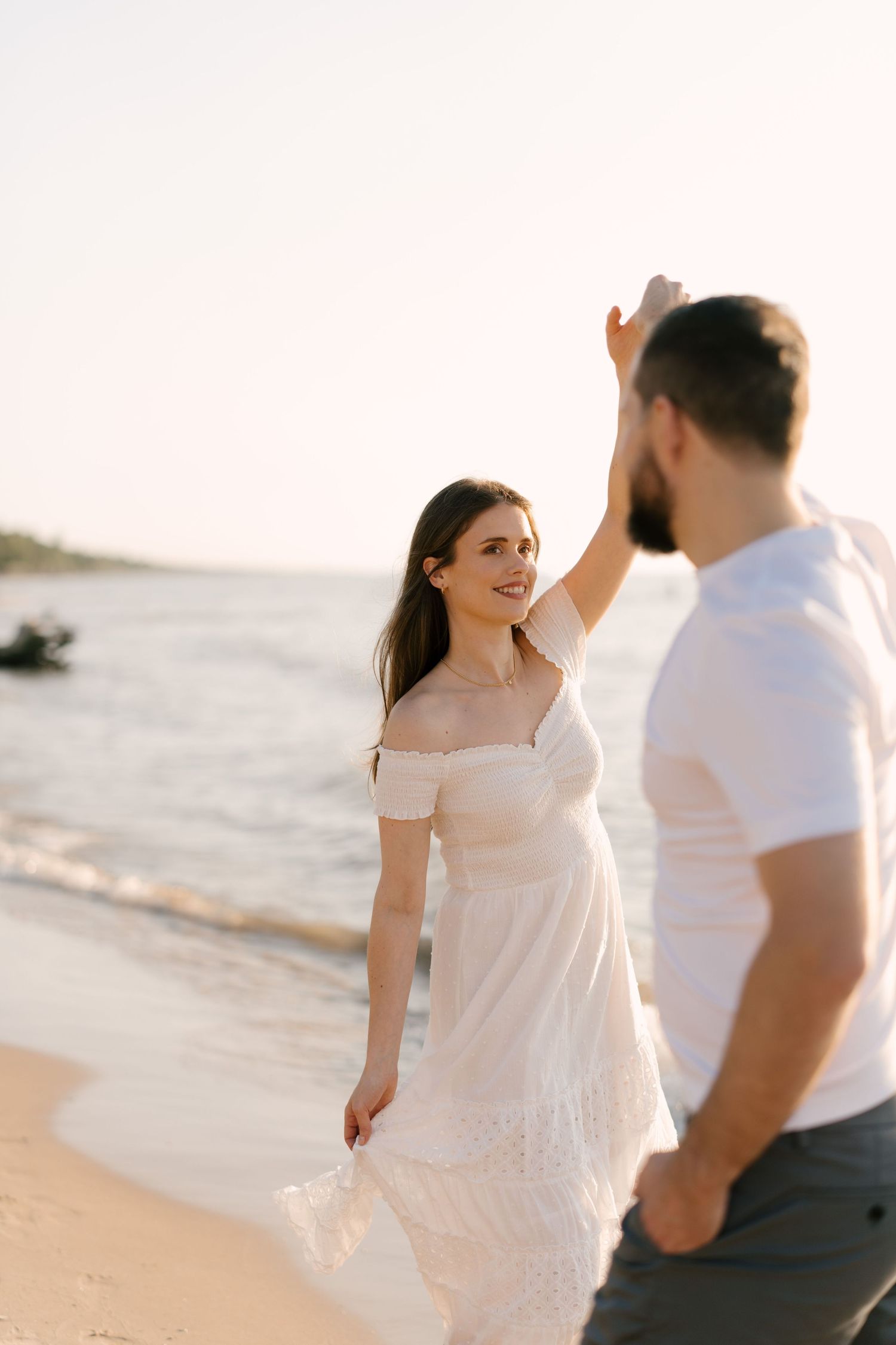 Woman in white off-shoulder dress dancing with partner on a sunlit sandy beach at golden hour.