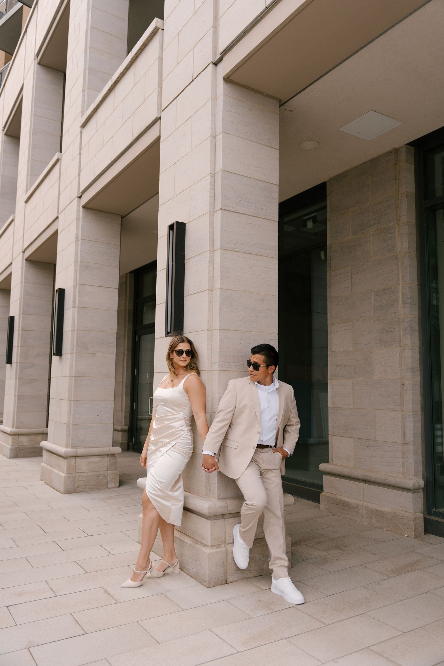 Elegant couple in matching beige outfits holding hands against stone columns of a modern building.
