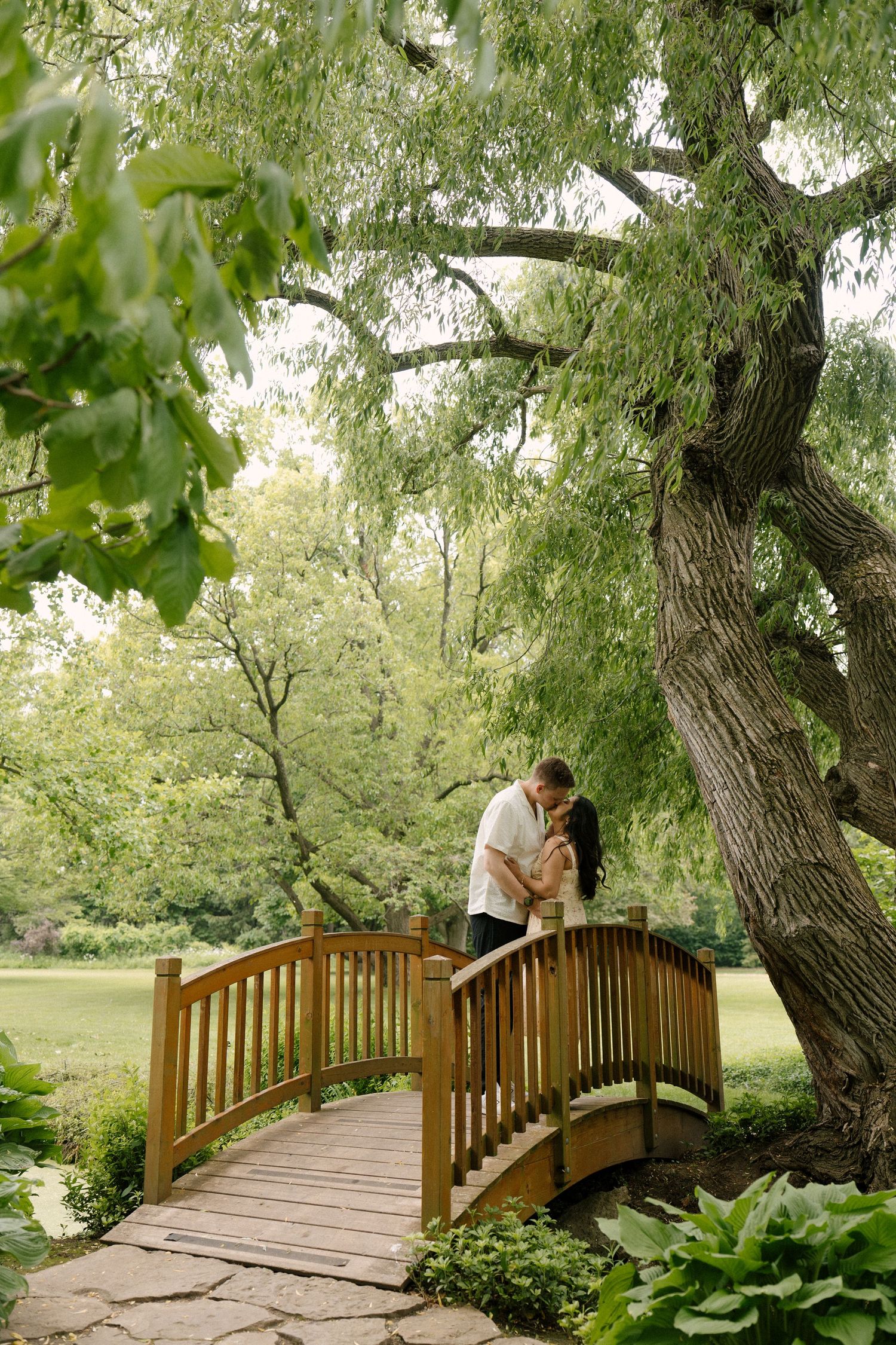 Couple kissing on a wooden arched bridge surrounded by lush green trees in a serene garden.