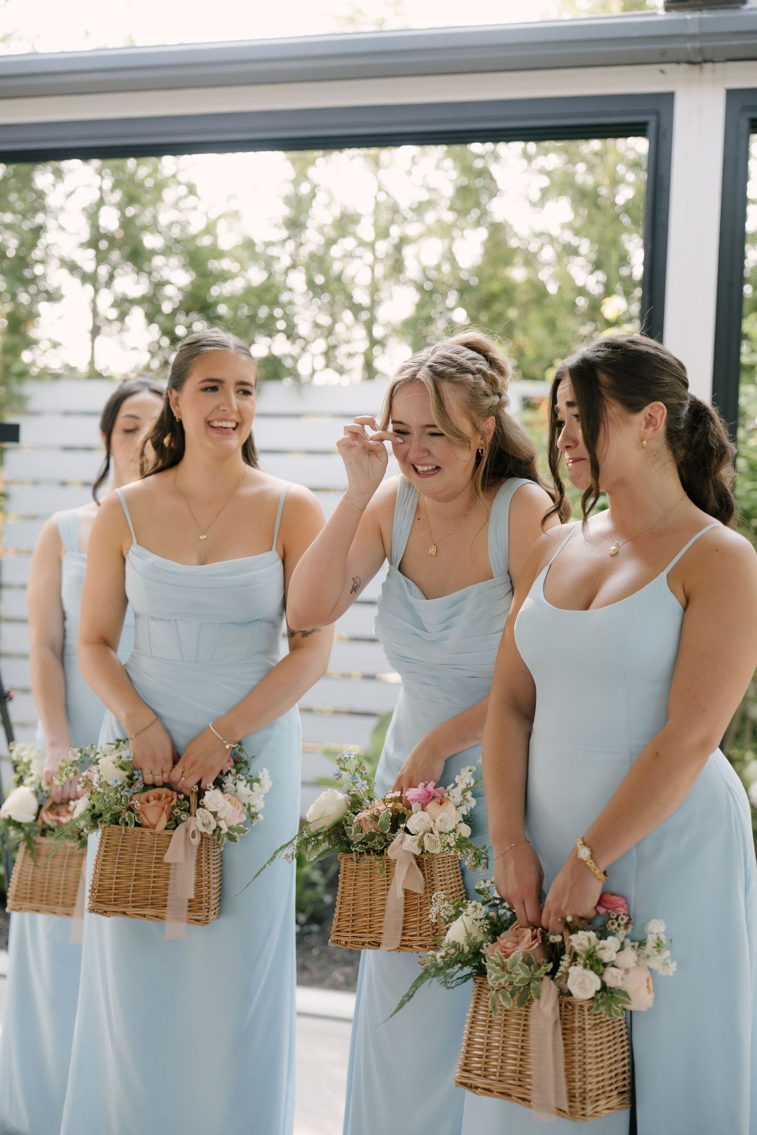 Four bridesmaids in light blue spaghetti-strap dresses laughing and holding wicker basket bouquets at Whistle Bear wedding.