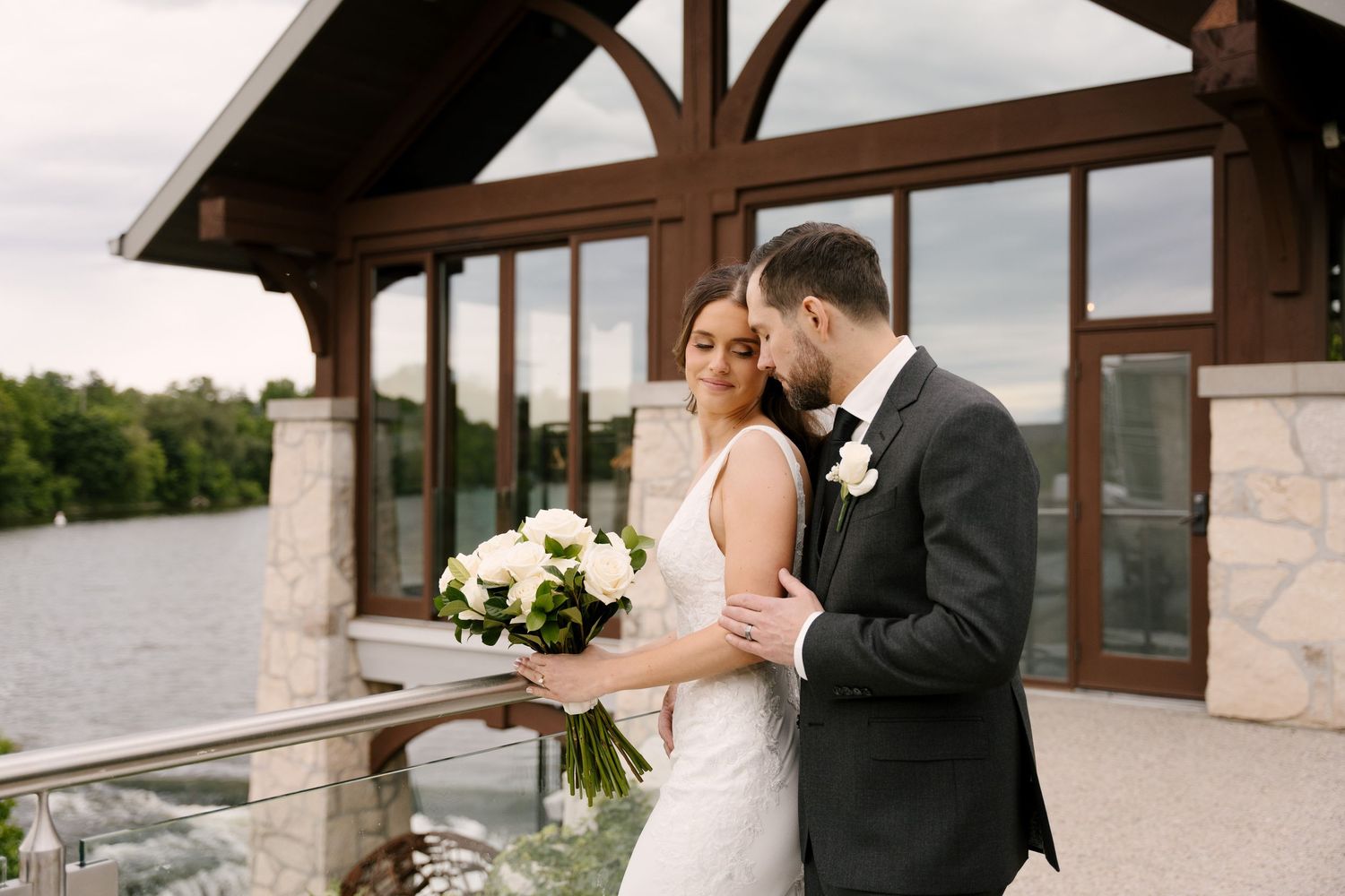 Bride and groom sharing a tender moment on a riverside Cambridge Mill venue balcony with white rose bouquet.