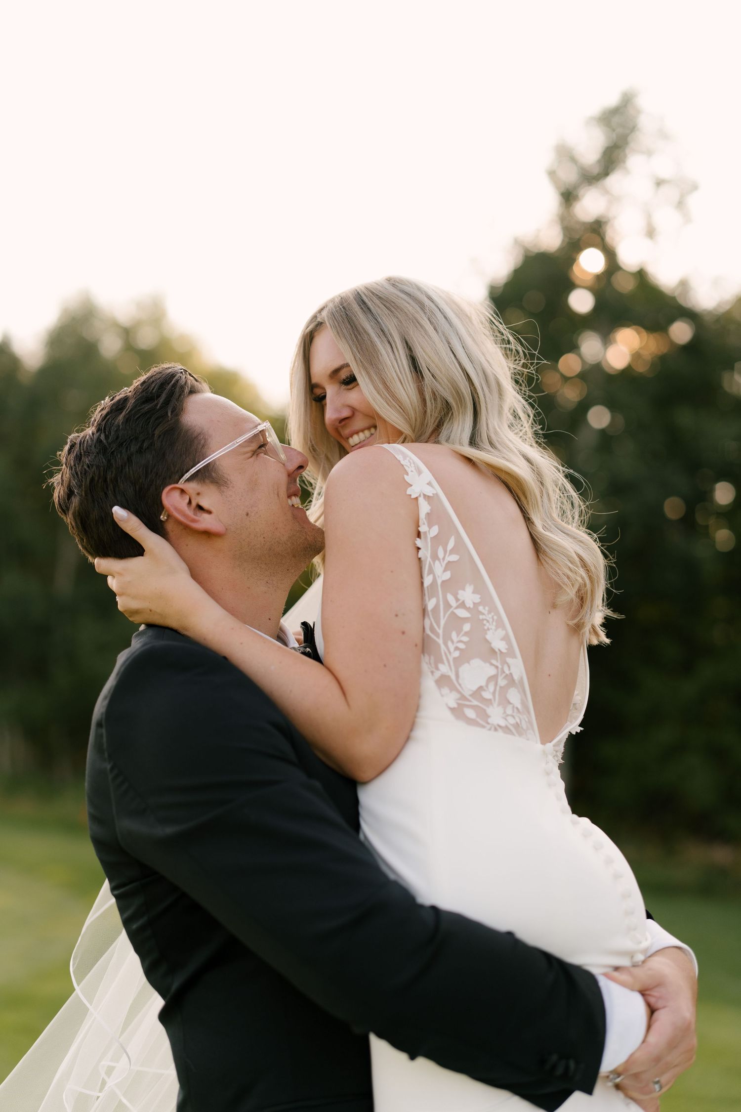 Groom in black tuxedo embracing and kissing smiling bride in backless lace wedding gown.