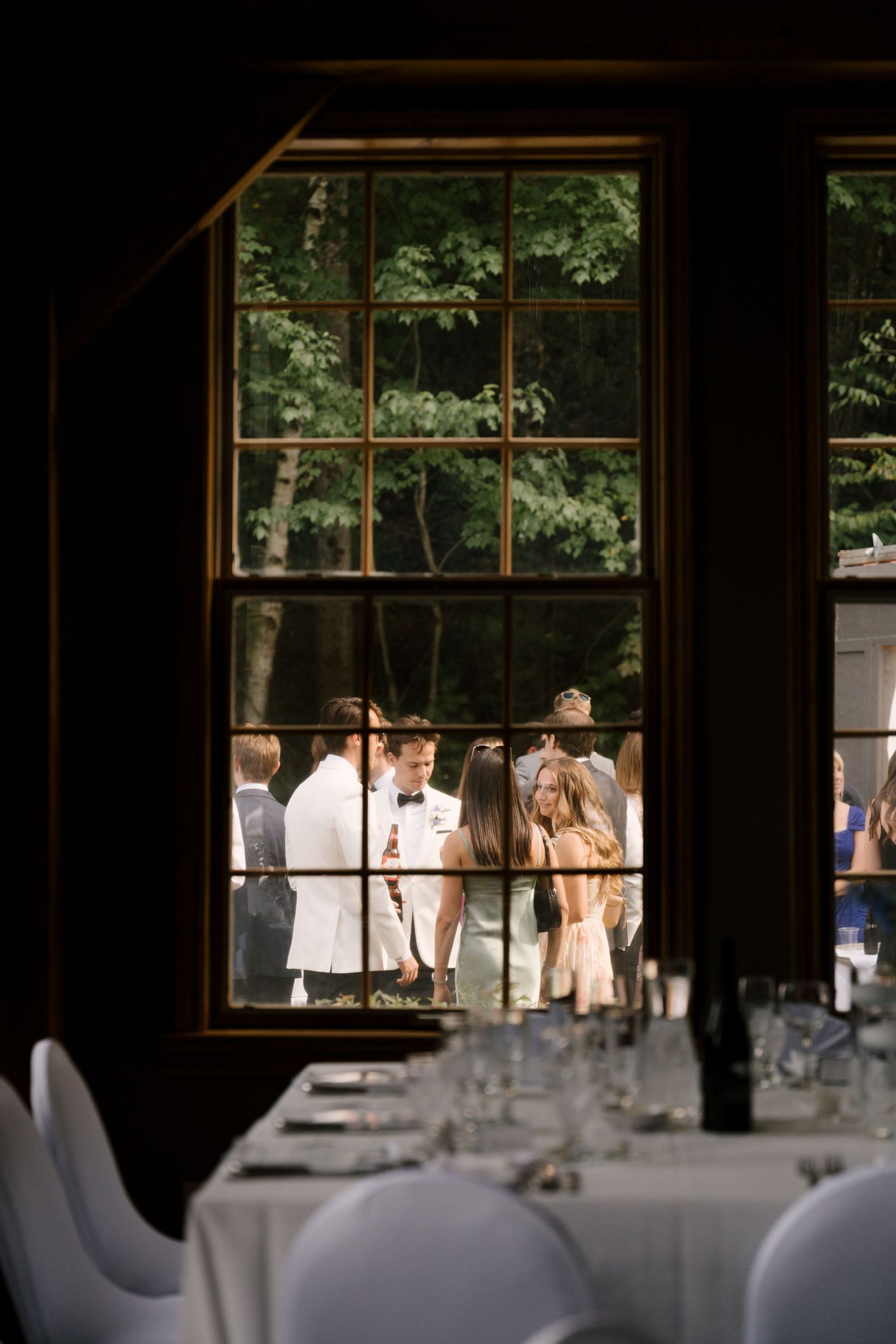 Wedding reception venue interior with set tables viewed through large window with guests mingling outside.