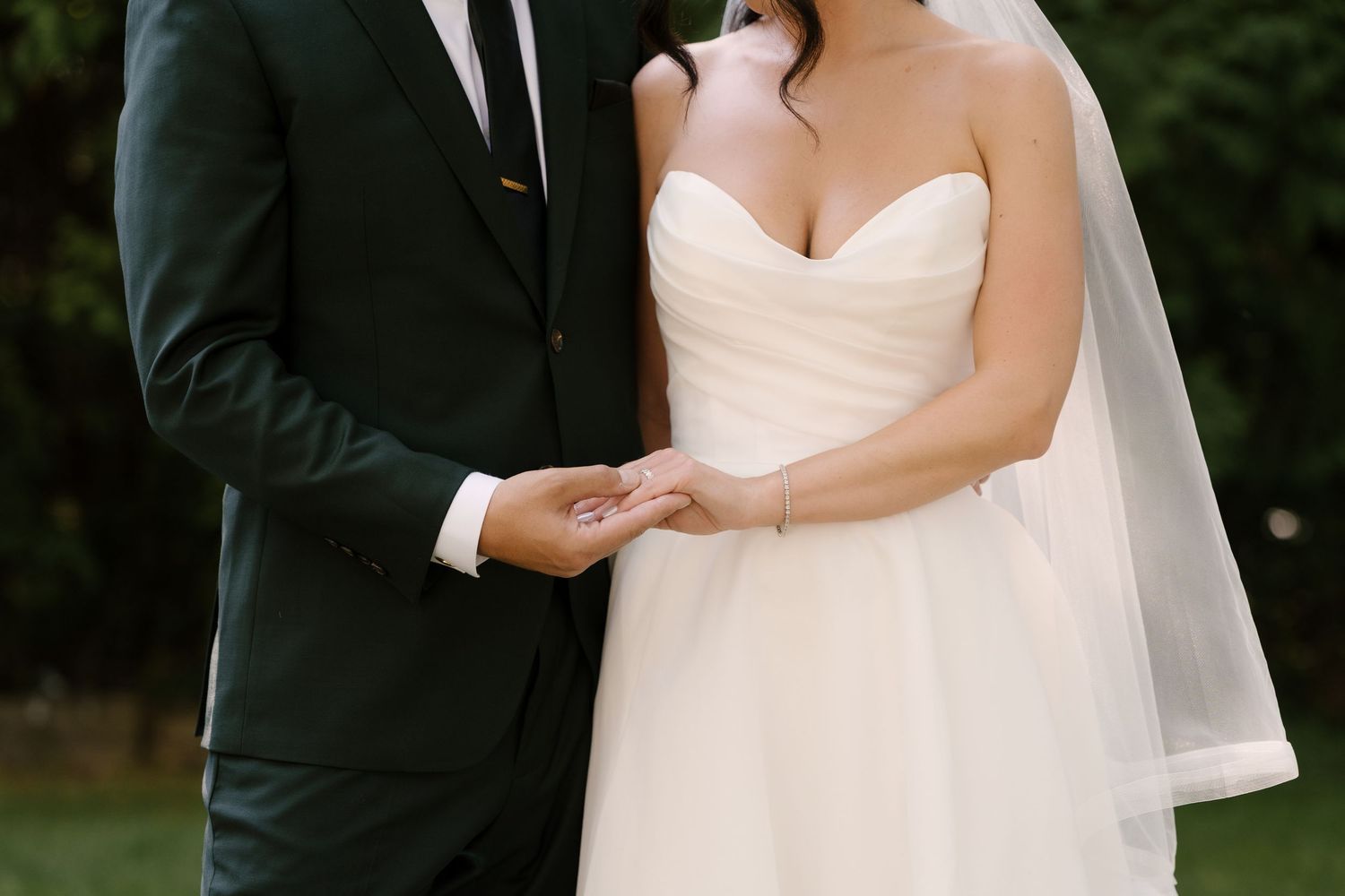 A bride in a strapless white gown and veil holds hands with a groom in a dark suit outdoors.