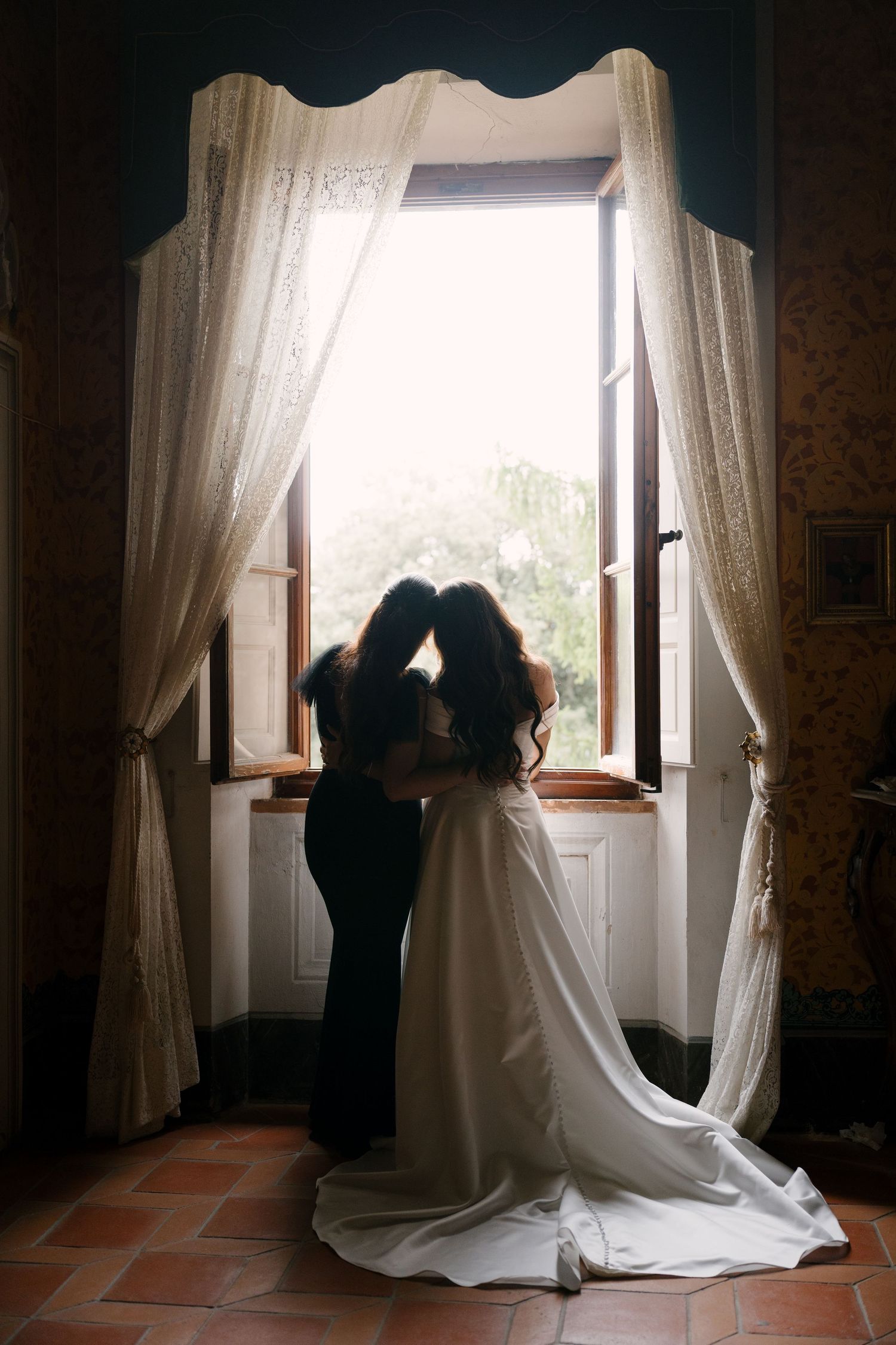 Bride in white gown and bridesmaid in black dress share intimate moment by sunlit window with lace curtains.