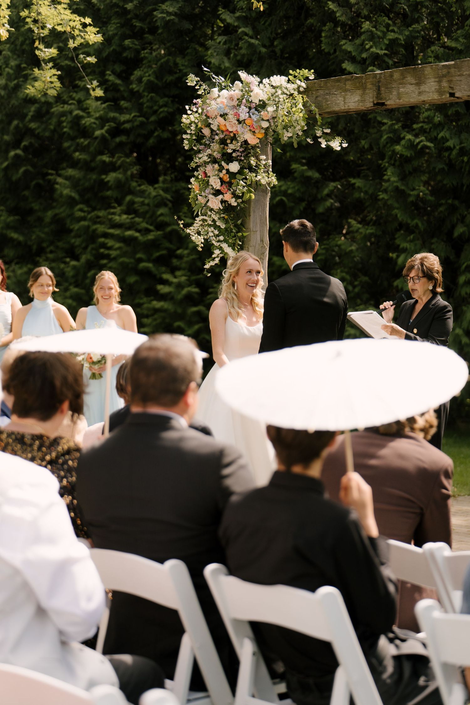Outdoor wedding ceremony with smiling bride at floral arch, guests seated in white chairs, bridesmaids in blue dresses nearby.