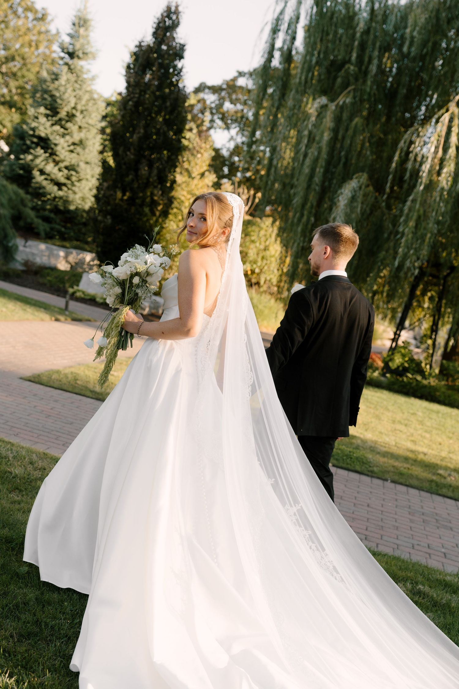 Elegant bride in backless white dress with cathedral veil strolls with groom in scenic Pillar and Post garden setting.