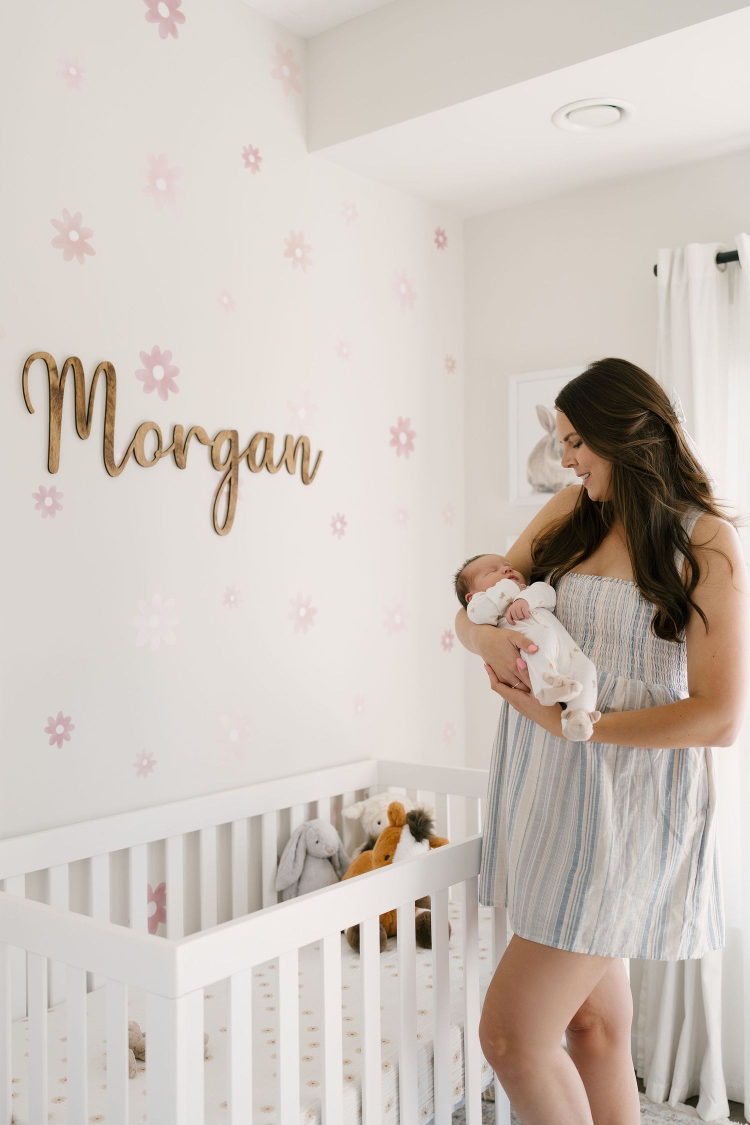 Smiling brunette mother holding newborn baby girl in floral nursery with Morgan name sign.