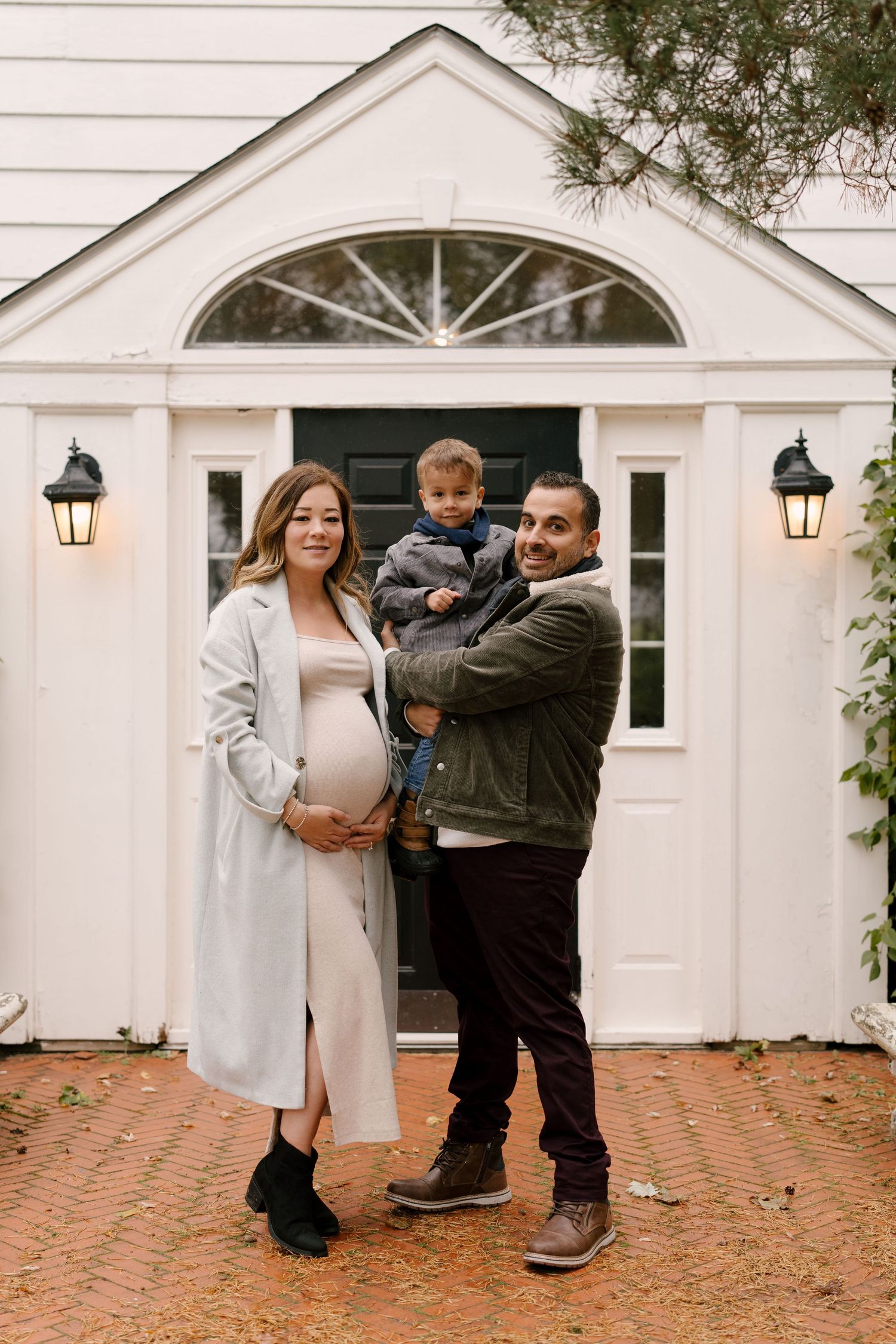 Pregnant woman and husband holding toddler posing in front of white colonial-style home.