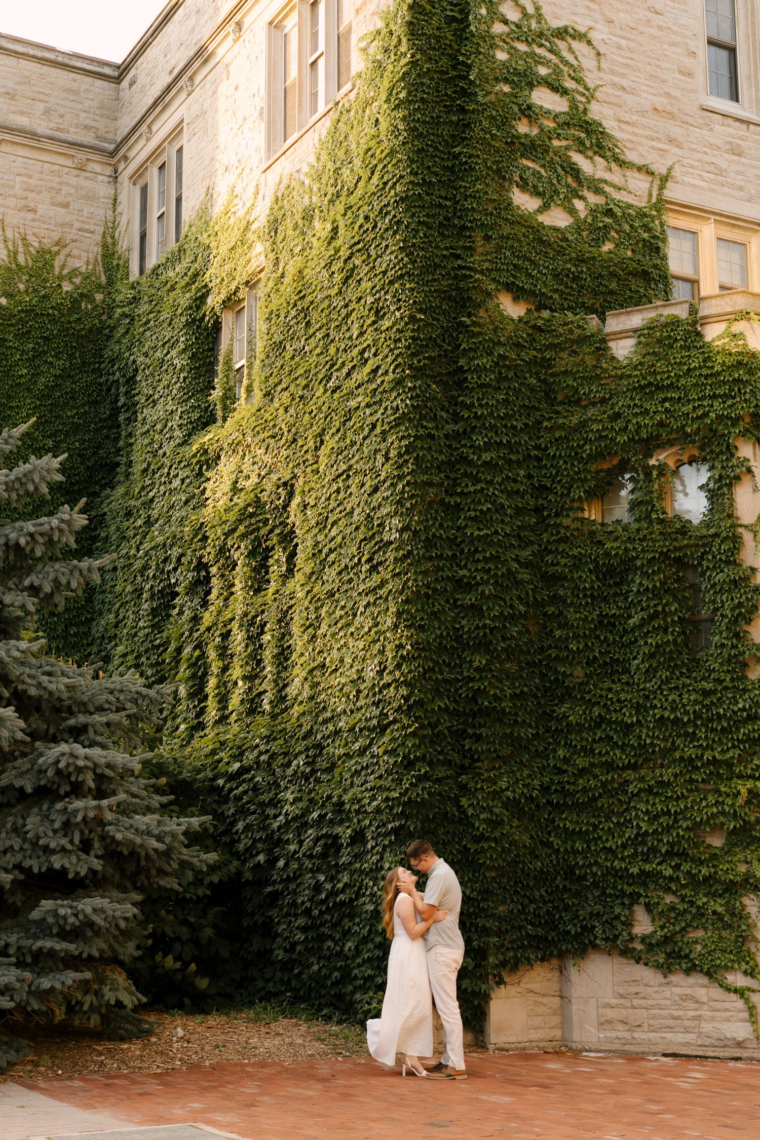 Couple embracing and kissing before a stunning ivy-covered stone building in warm golden light.