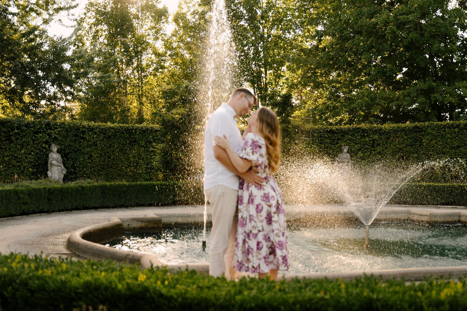 Romantic couple kissing near ornate garden fountain surrounded by lush hedges at golden hour.