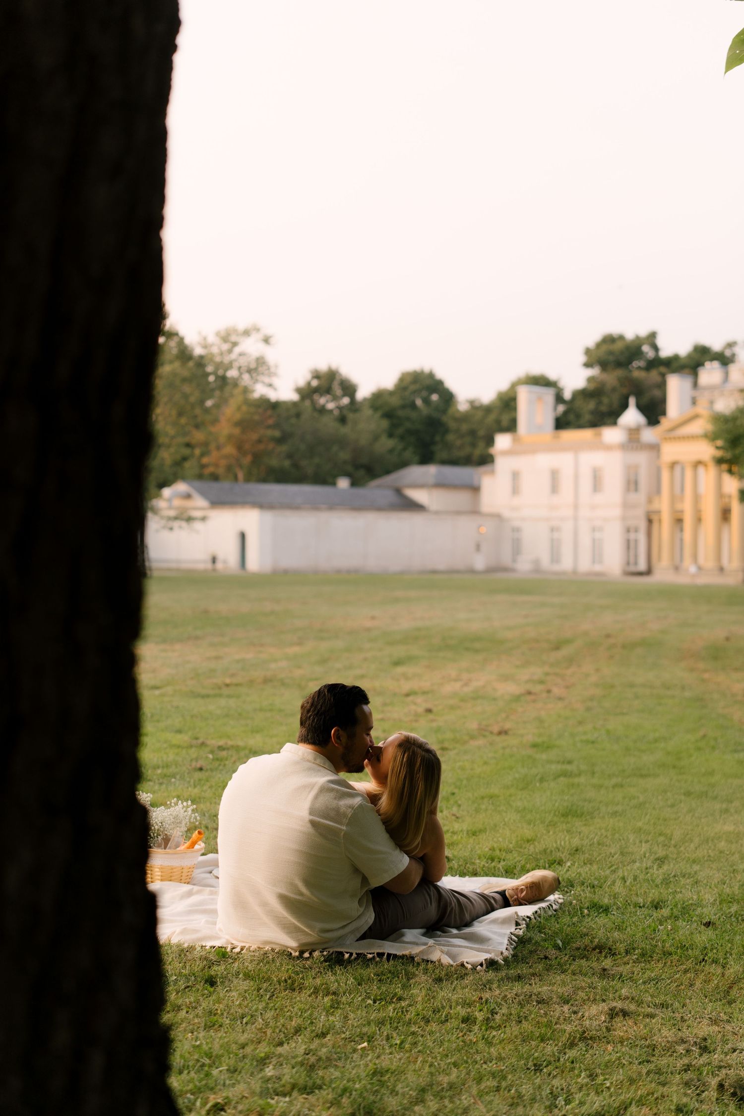 Couple kissing on a picnic blanket on a green lawn with a historic white mansion in the background.