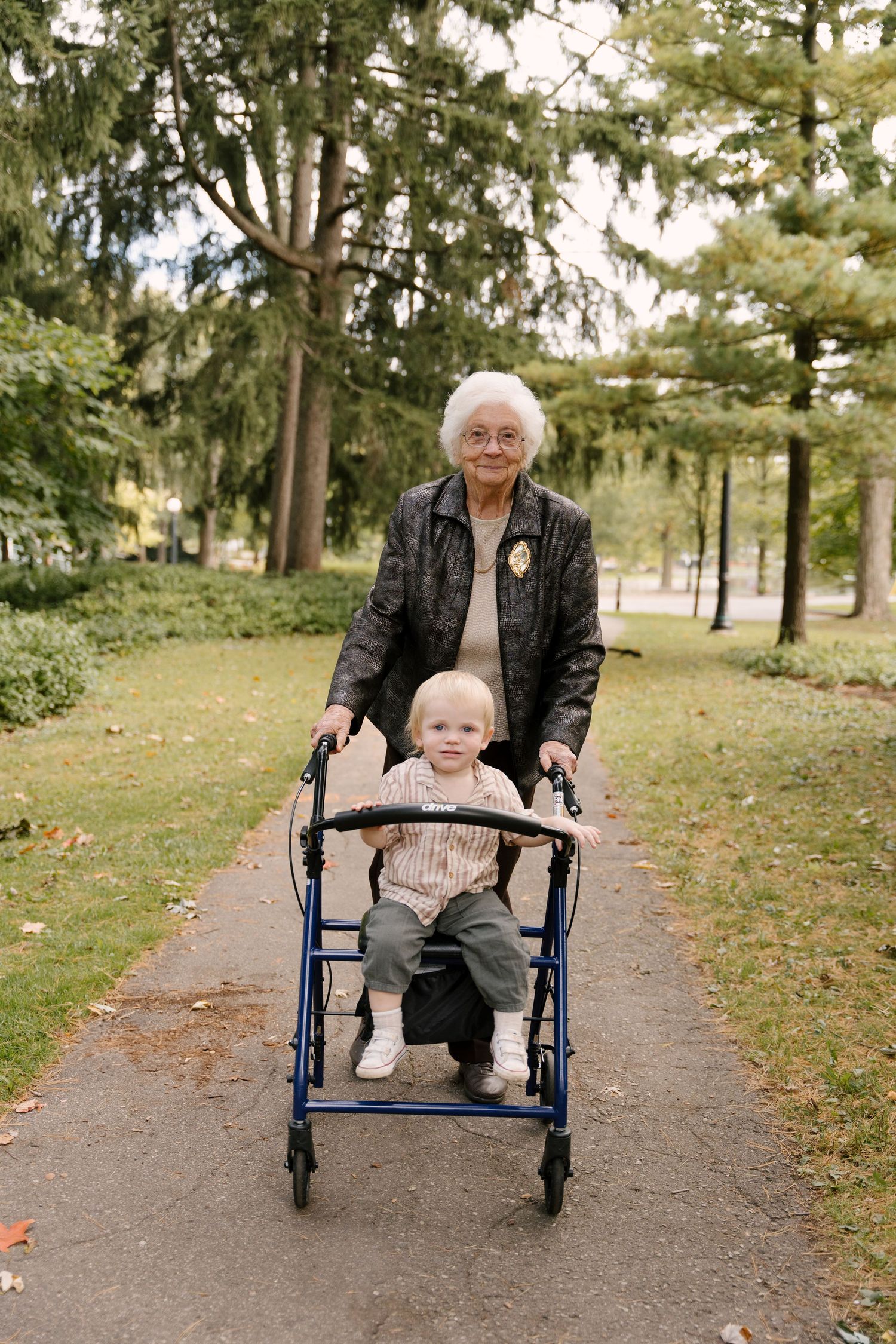 Elderly grandmother pushing baby in walker along a scenic green park pathway.