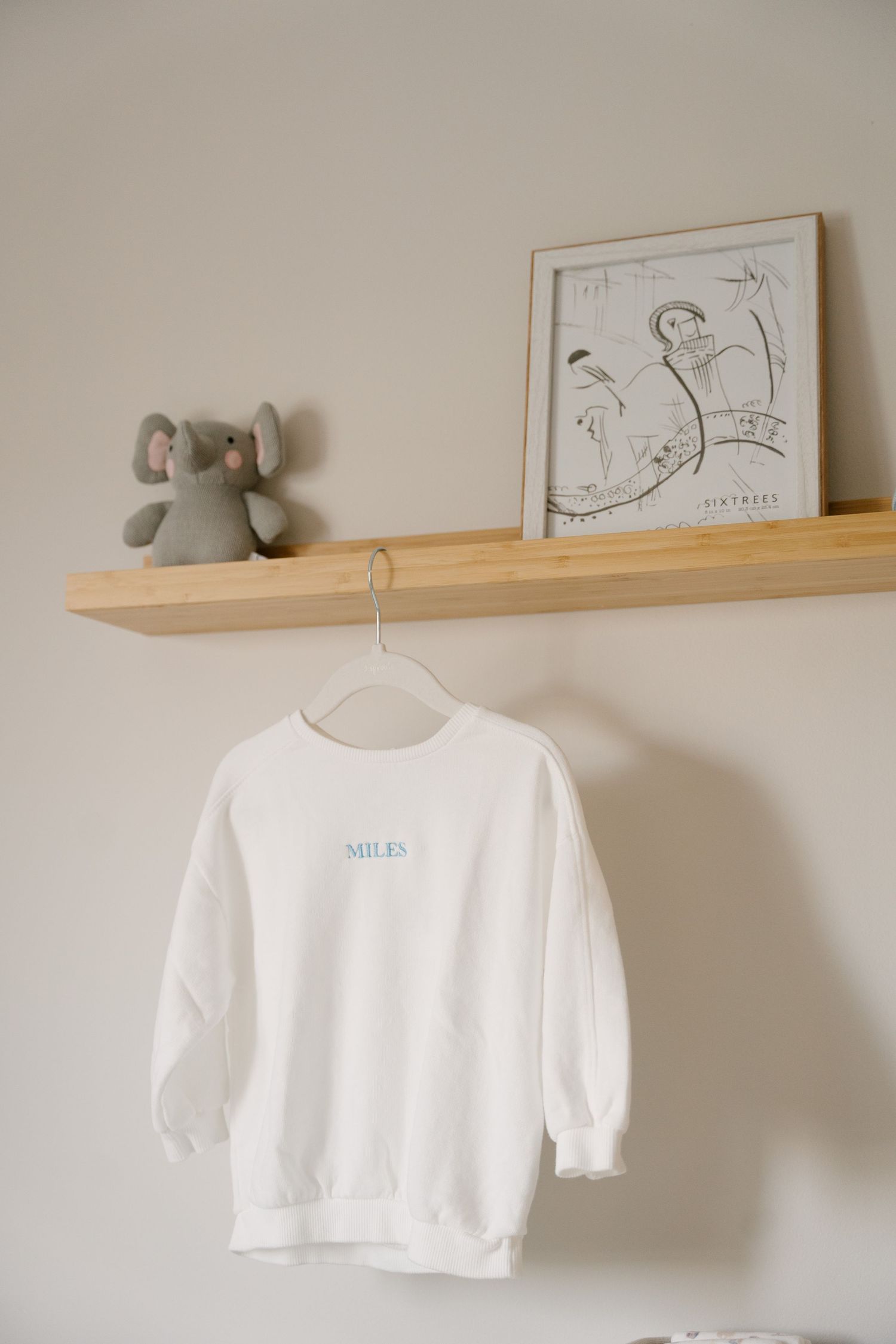 White personalized sweatshirt with name Miles hanging on nursery shelf with elephant toy and framed art.